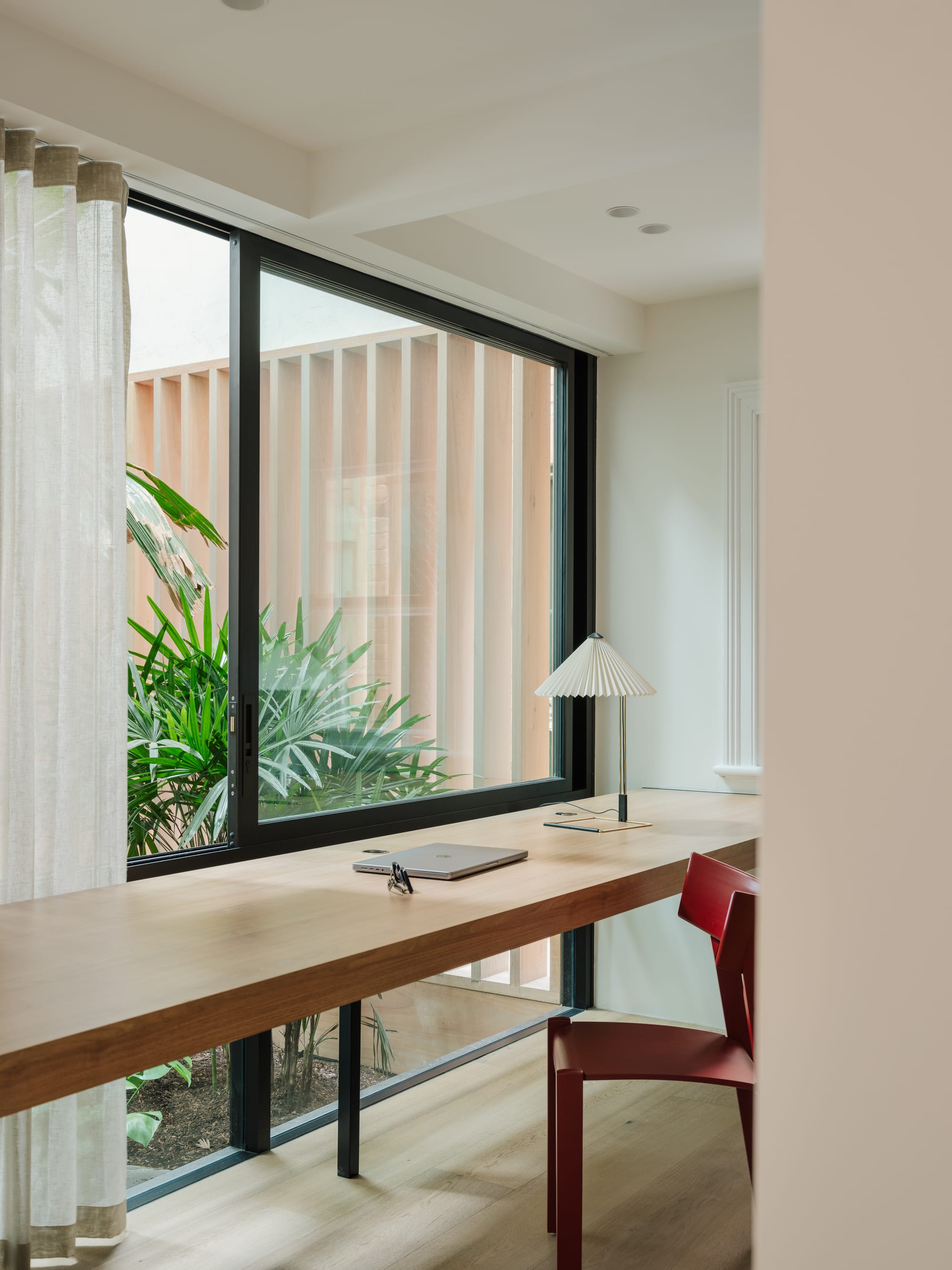 Petersham House by Cateaux Architects with CHROFI. Photography by Clinton Weaver. Floating timber bench in study with red chair in front of full length window overlooking green tropical plants. 