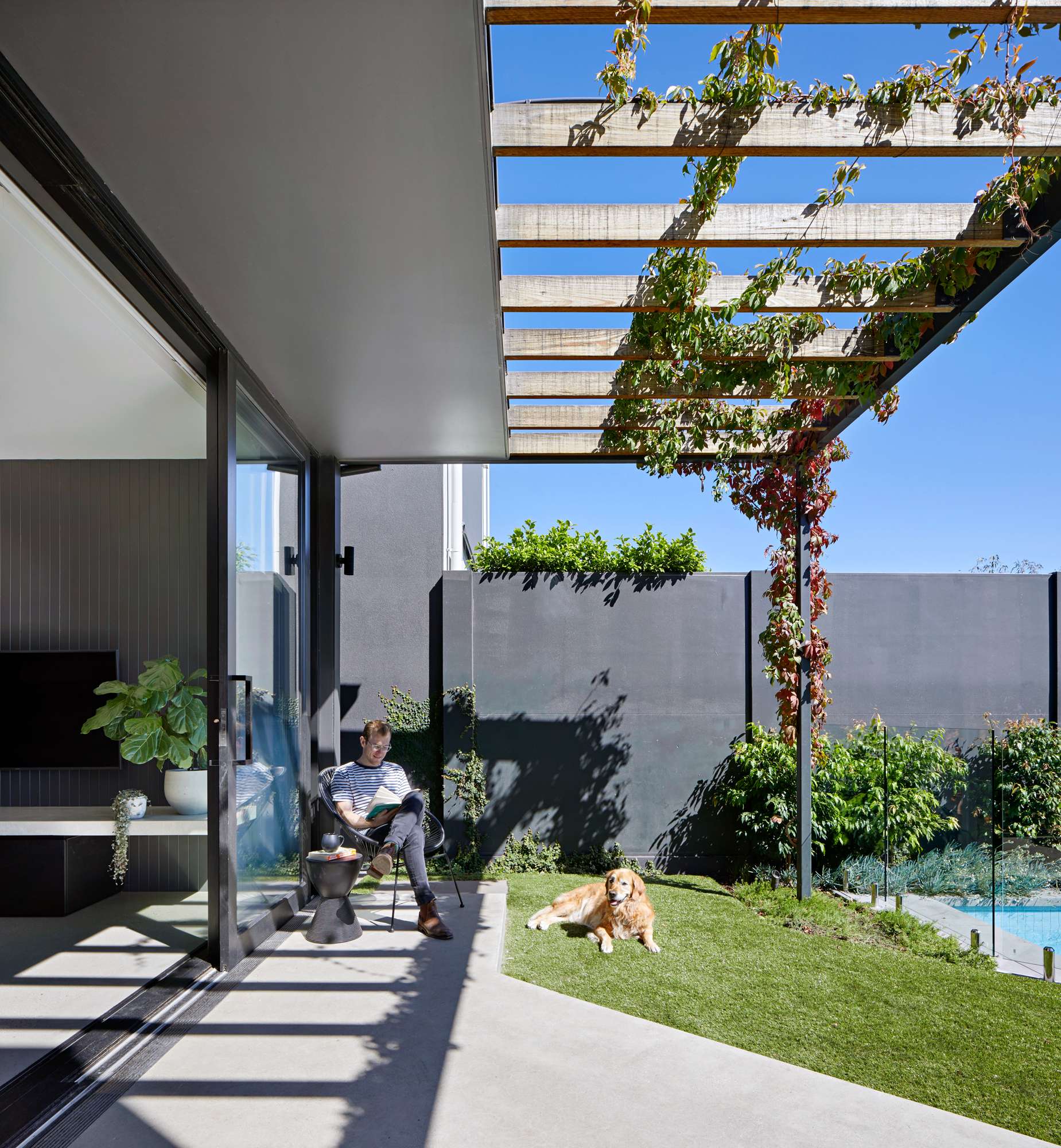 Fitzroy North House by BENT Architecture. Photography by Tatjana Plitt. Outdoor patio with large sliding black framed door connecting garden with living space, green grass, a pool to the right and a man reading a book with his dog.