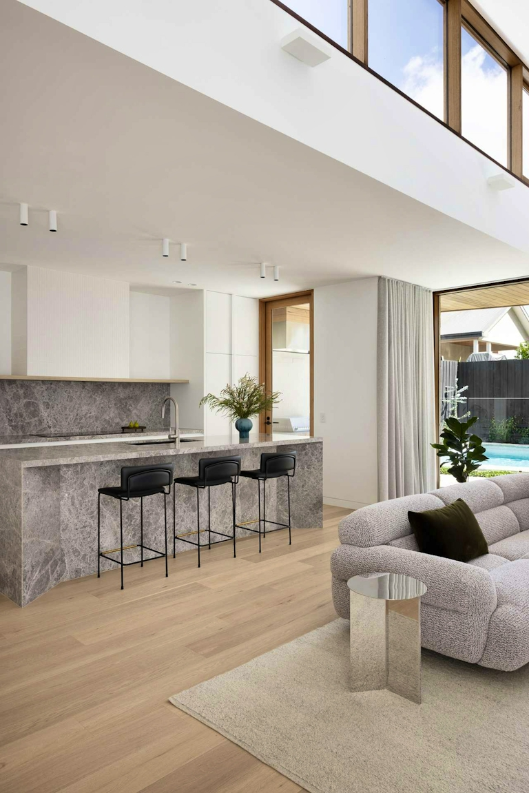 Bastings House by FURNISHD. Photography by Dylan James. Open plan living and kitchen space with marbled stone countertops and splashback, a modular grey lounge, light timber floors and a pool visible through the full height glass windows. 