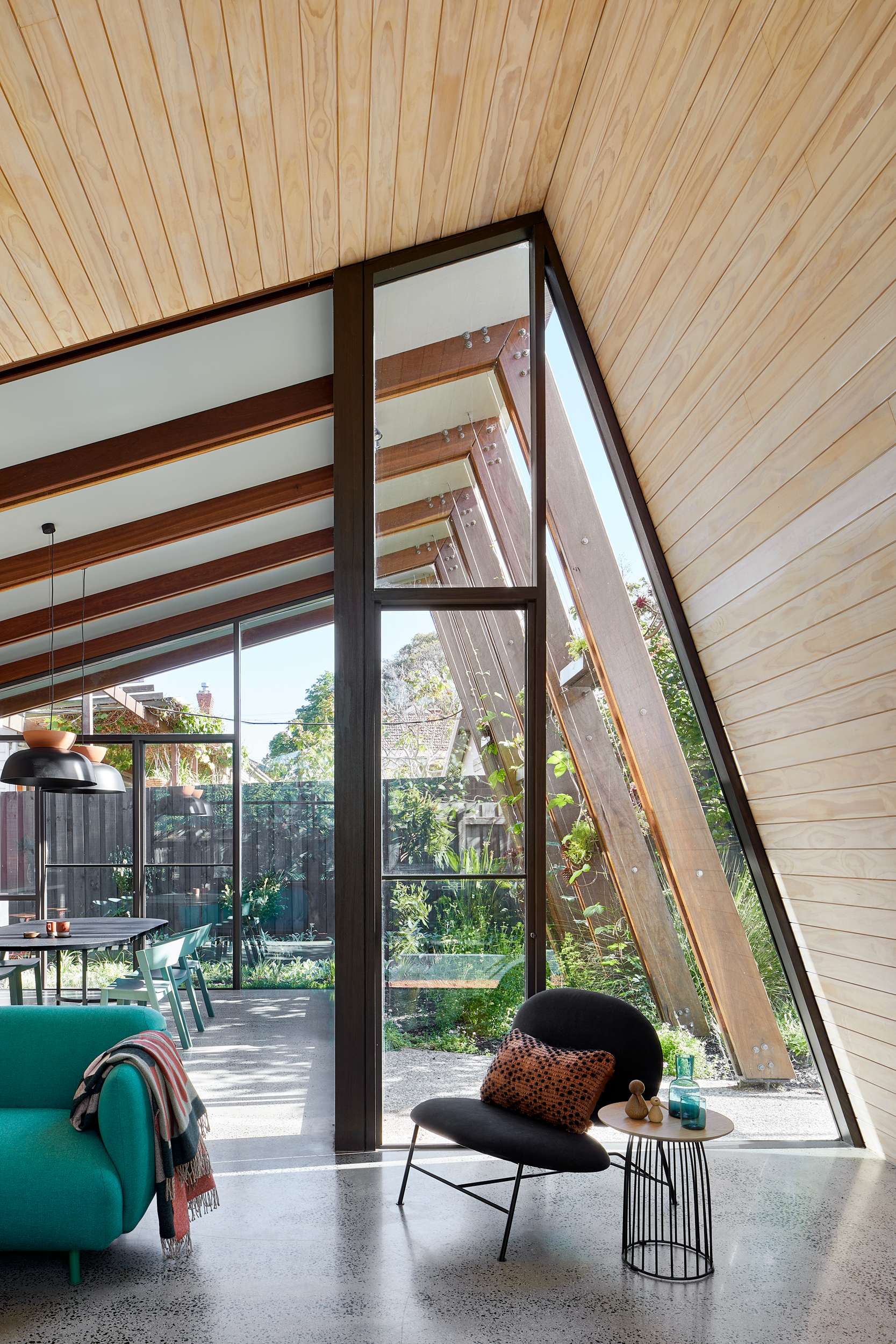Bent Annexe II by BENT Architecture. Photography by Tatjana Plitt. Concrete flooring in open plan living and dining area with pitched timber clad ceilings and angled glass windows, opening onto lush garden. 