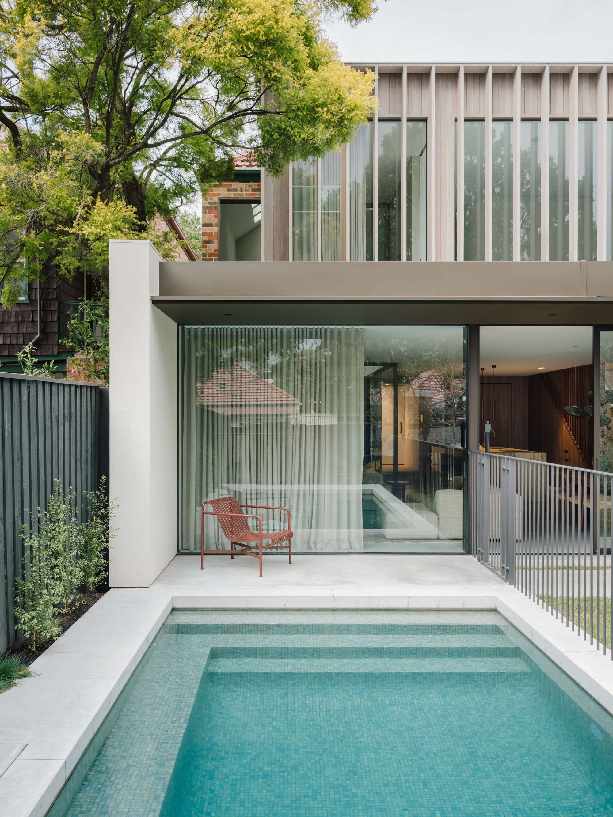 Petersham by Cateaux Architects. Photography by Clinton Weaver. Rear facade of double storey home with a tiled mosaic pool, white tiles patio, grey fencing and full height windows looking into interior living and kitchen space.