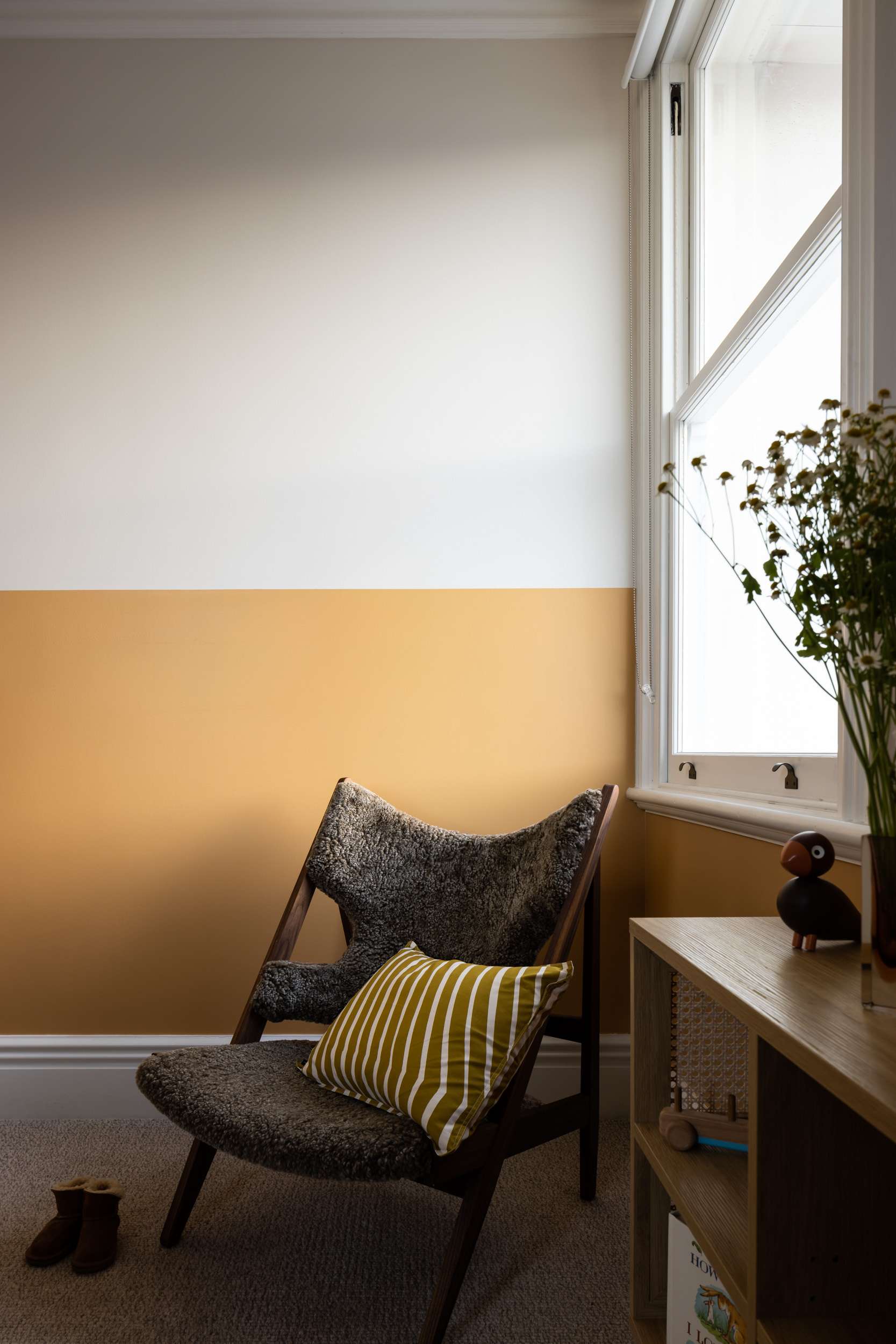 Dappled Terrace House by Marker Architecture. Photography by Simon Whitbread. Nursey with half white, half yellow walls, a low-slung armchair and a bookshelf storage unit with toys and flowers. 