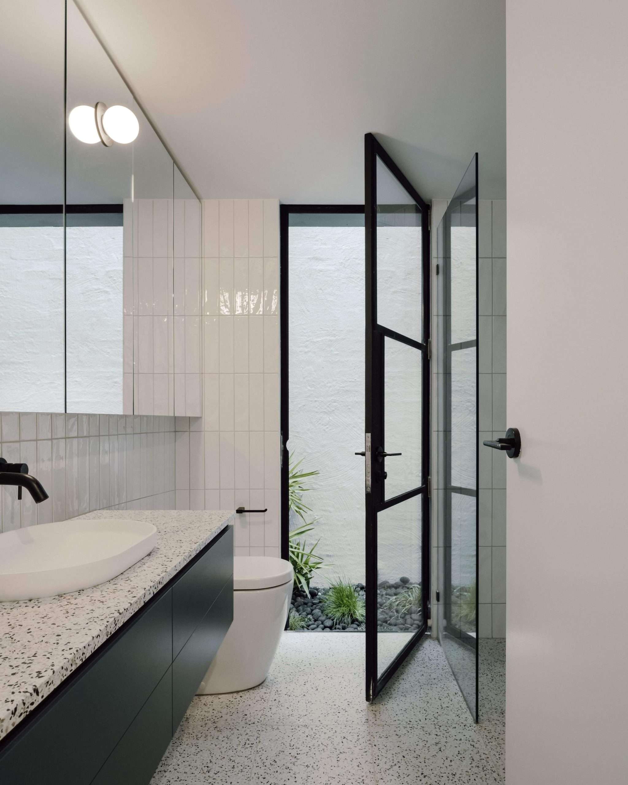 Richmond Terrace by Eckersley Architects. Photography by Dan Preston. Bathroom with large black framed glass door opening onto outdoor gardening, dark green floating cabinetry with terrazzo countertops and white tiled walls.