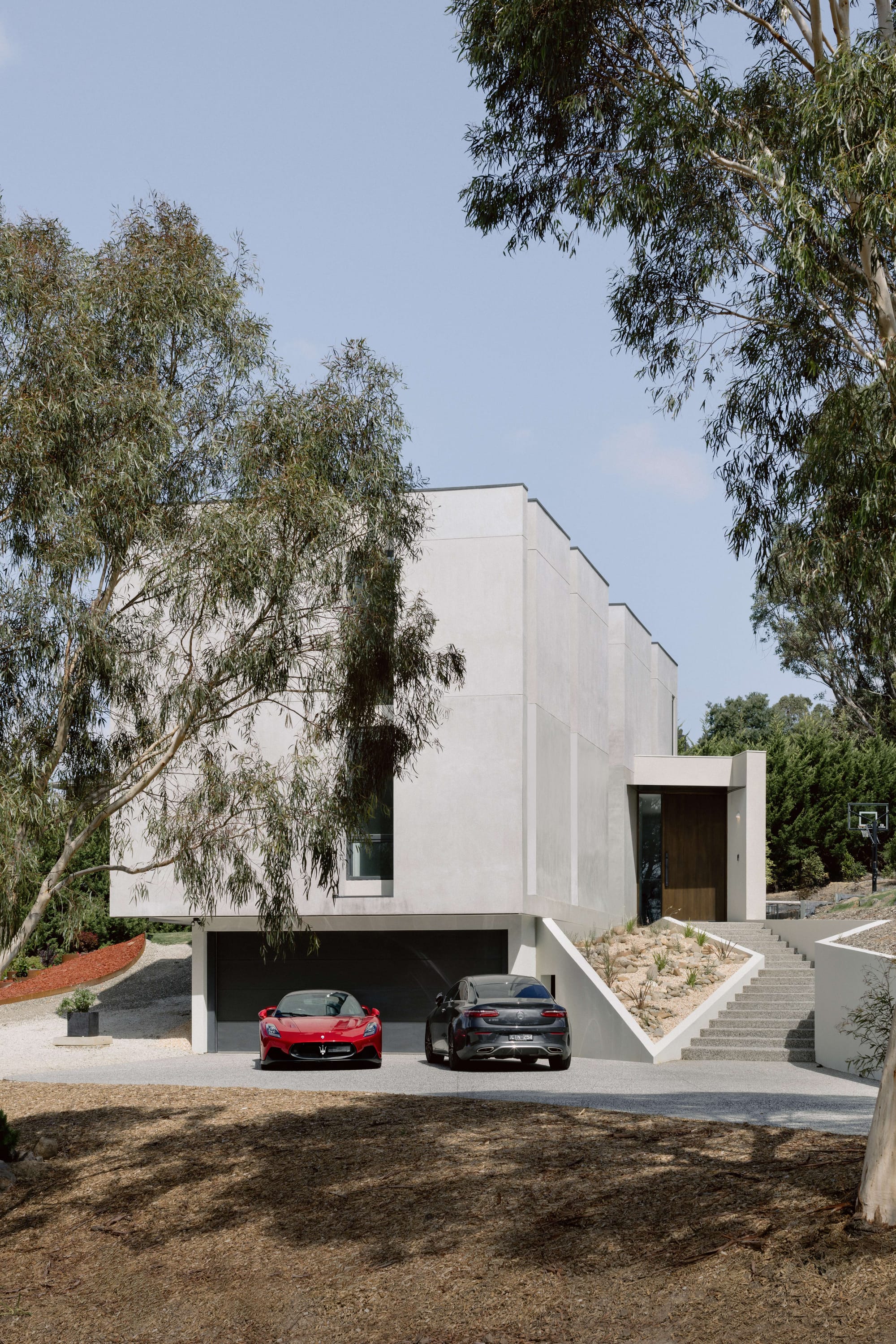 House On The Hill by Enclave Architects. Photography by Elise Scott. Front facade of multi storey, modern home with a concrete exterior, large sweeping steps to the left and a red sportscar and black SUV parked on poured concrete driveway.