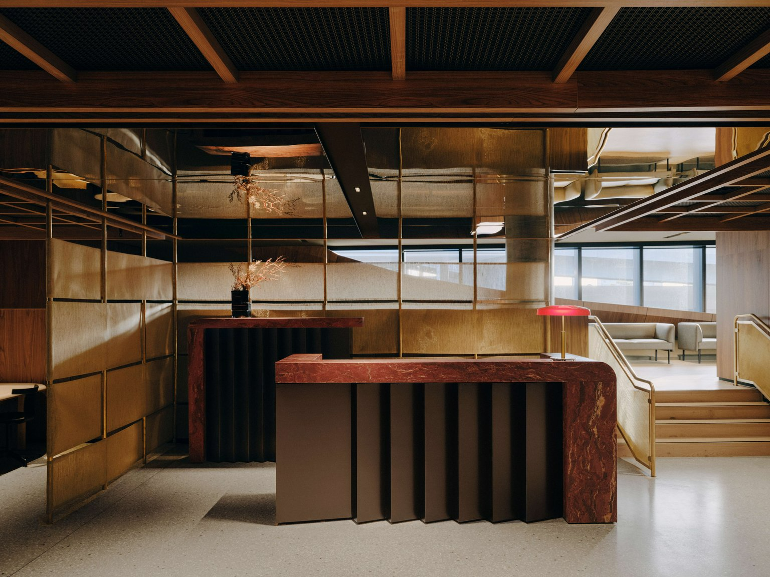 Aerofoil Workspace Melbourne Airport by FK. Photography by Tom Ross. Interior reception space with gold chrome paneled walls, a dark brown and red marble reception desk and exposed terracotta overhead grates. 