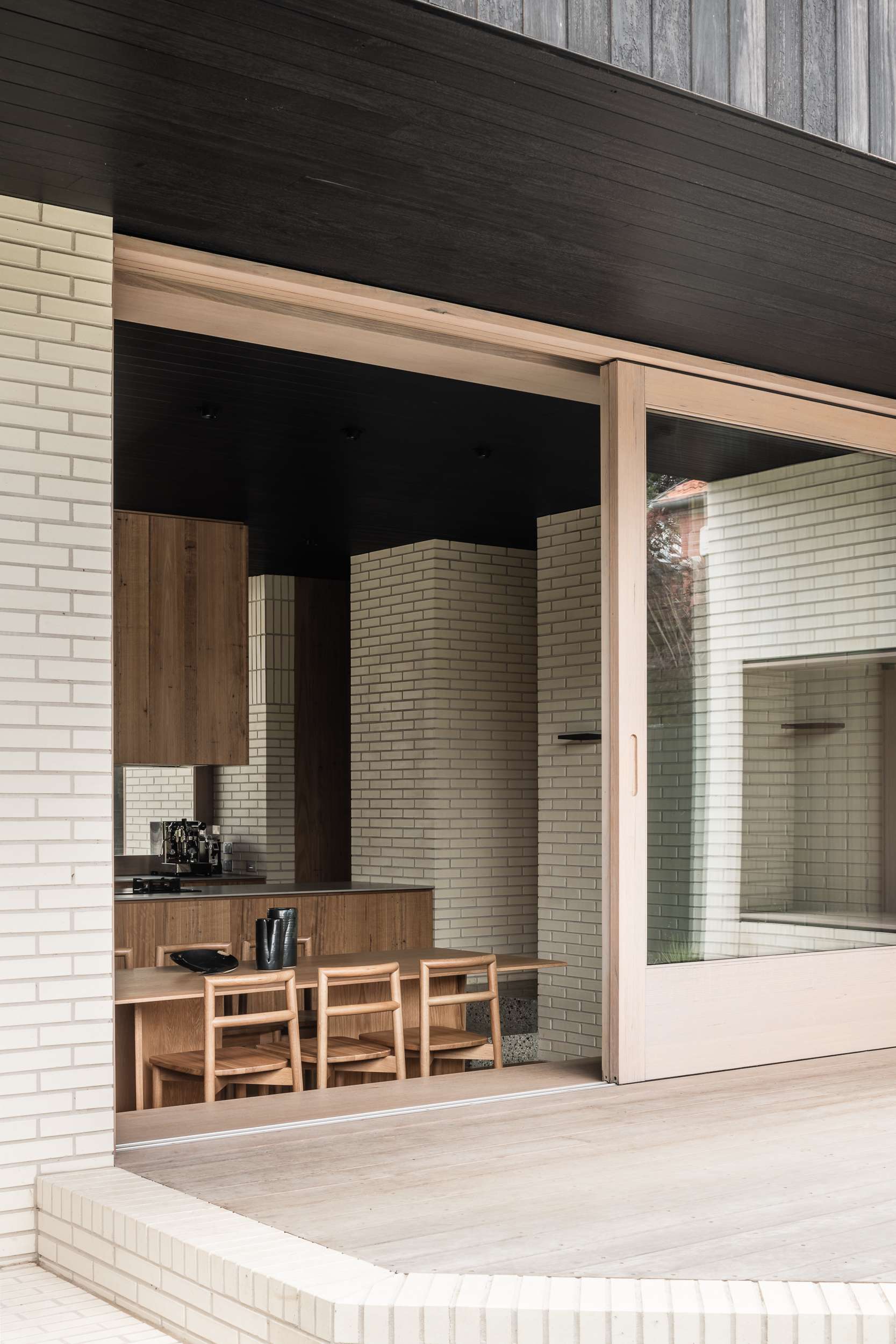 Ha Ha Haus by FIGR Architecture. Photography by Tom Blachford. Rear facade of home with pale timber decking and brick walls, stepping down into an open plan kitchen and dining space with warm timber cabinetry and furnishings. 