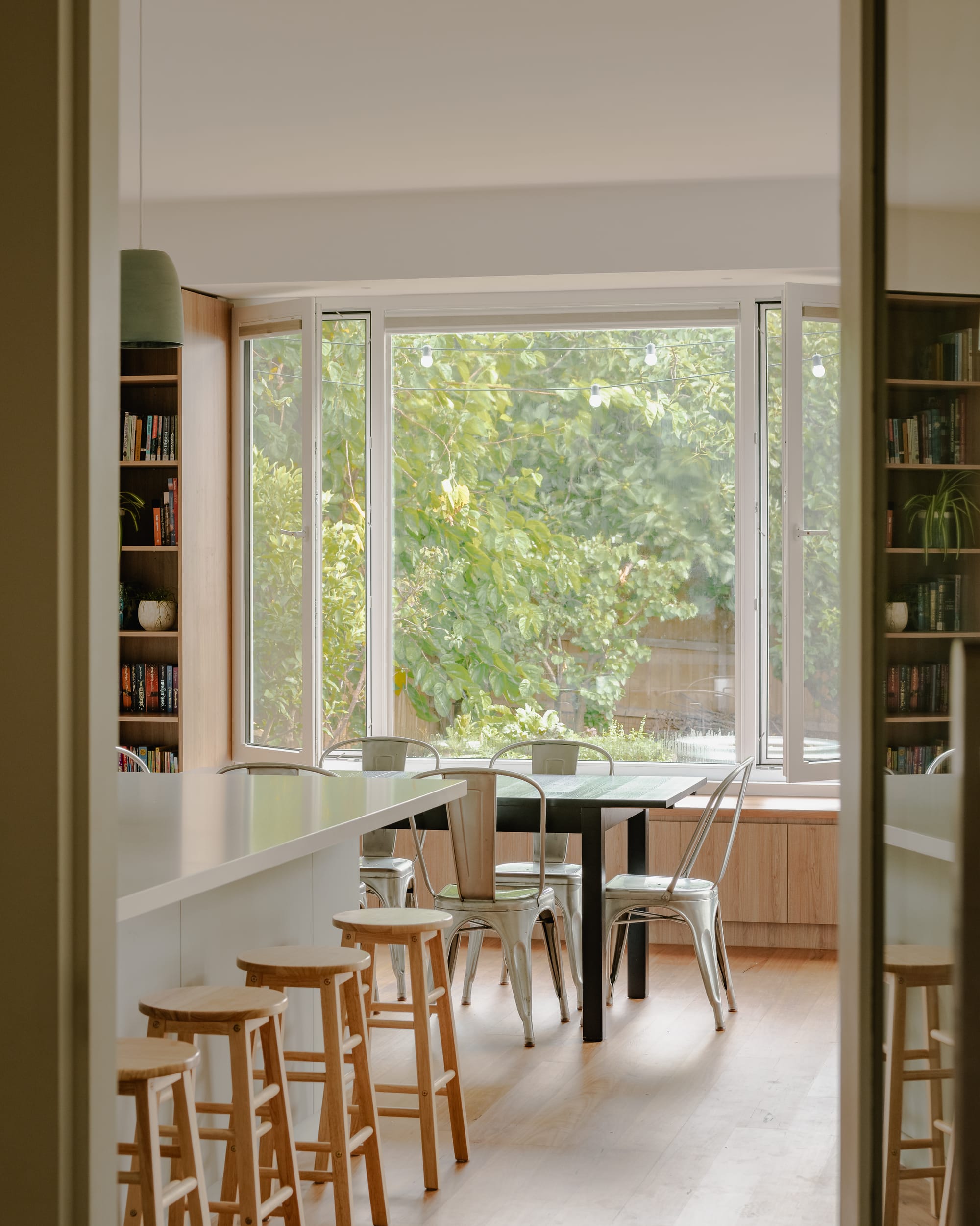 House in Preston by Ashley O'Neill Architects. Photography by Victor Vieaux. Open plan kitchen and dining space with timber stools at island bench in the foreground, and a dark timber table with silver cafe-style chairs in foreground, positioned in front of a timber window seat and a window opening inwards and offering views over the garden. 