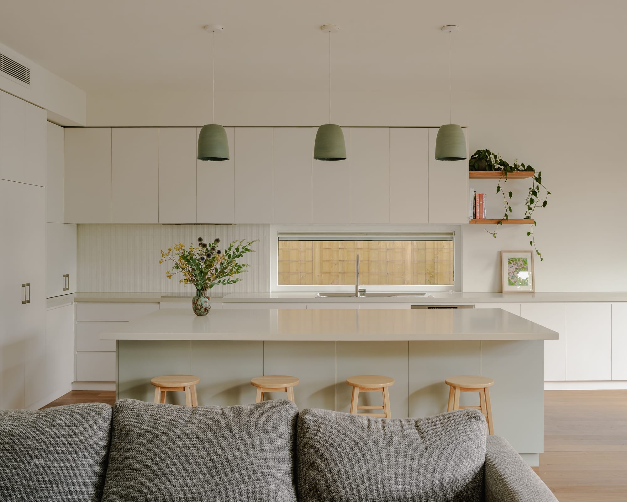 House in Preston by Ashley O'Neill Architects. Photography by Victor Vieaux. Kitchen with simple white cabinetry running the full length of the space, an island bench with timber stools and light green kickboard, hanging green pendant lighting and a grey couch in the foreground. 