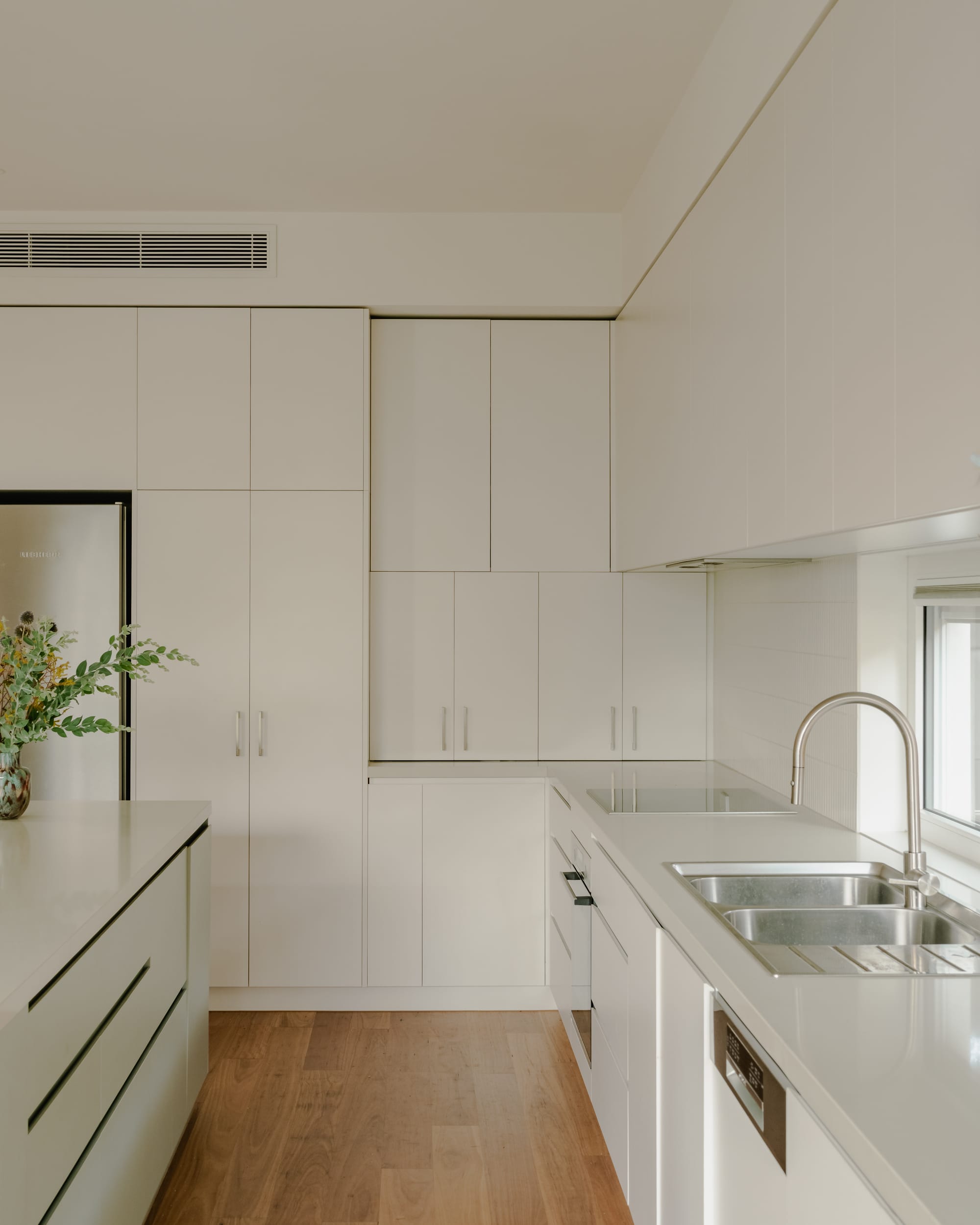 House in Preston by Ashley O'Neill Architects. Photography by Victor Vieaux. Kitchen with simple white cabinetry, timber floors, silver appliances and a vase of fern leaves on left hand side. 