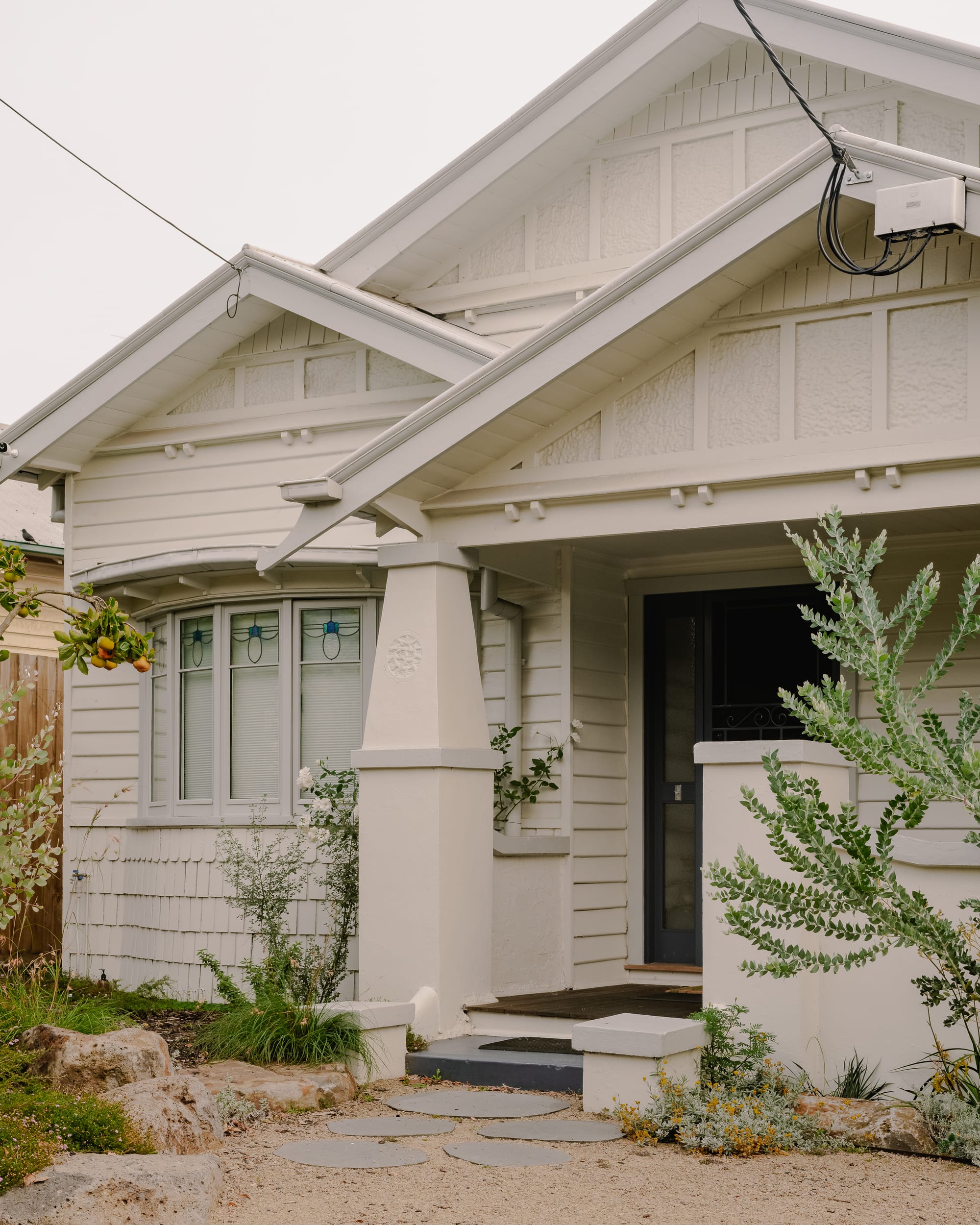 House in Preston by Ashley O'Neill Architects. Photography by Victor Vieaux. Front facade of bungalow-style home with timber cladding, stepped timber porch, leadlight windows and pitched roof, all finished in cream, off-white paint colour. 