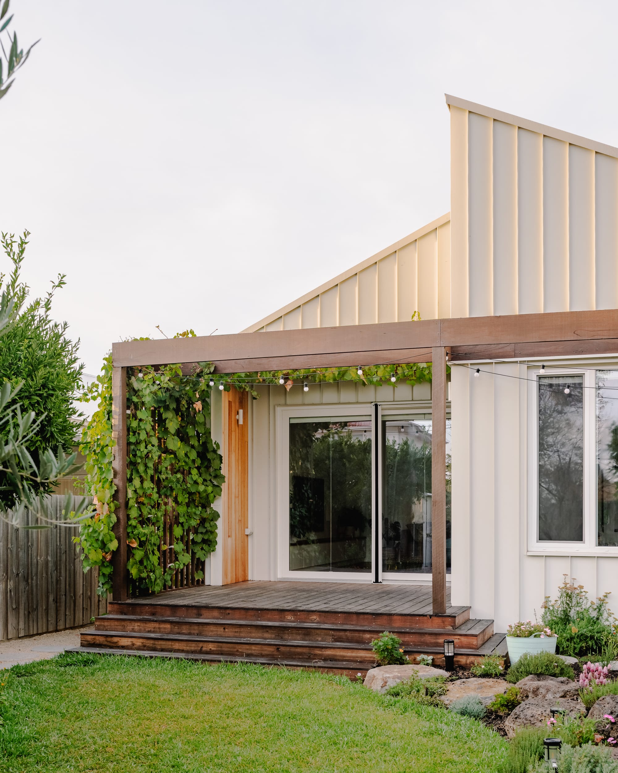 House in Preston by Ashley O'Neill Architects. Photography by Victor Vieaux. Rear facade of home with timber porch, pergola covered in vines, metal cladding on exterior, grass and a garden to the right which features rocks, plants and pink flowers. 