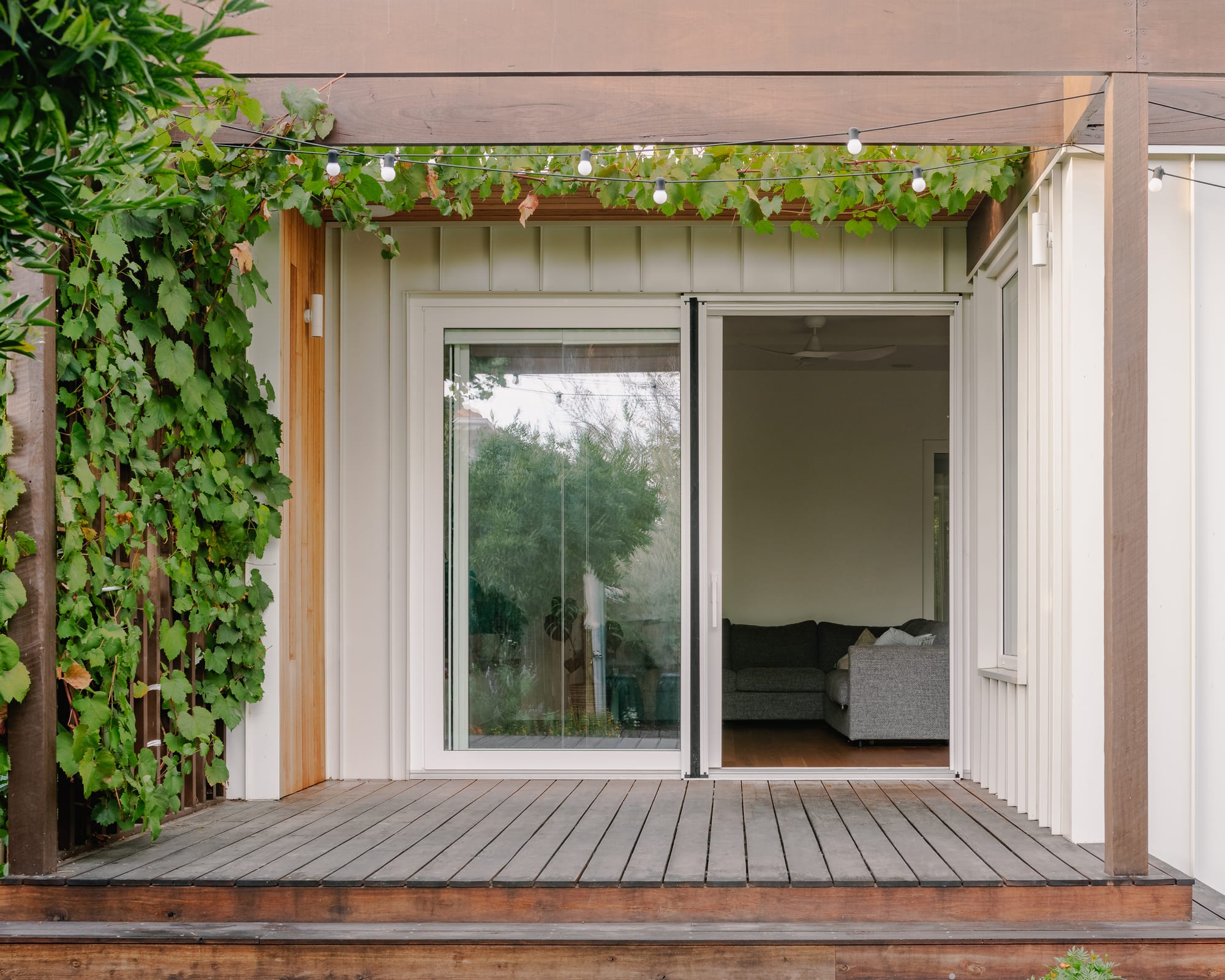 House in Preston by Ashley O'Neill Architects. Photography by Victor Vieaux. Tiber porch with a pergola covered in vines, with full height sliding glass doors connecting to an interior living space with a grey couch. 