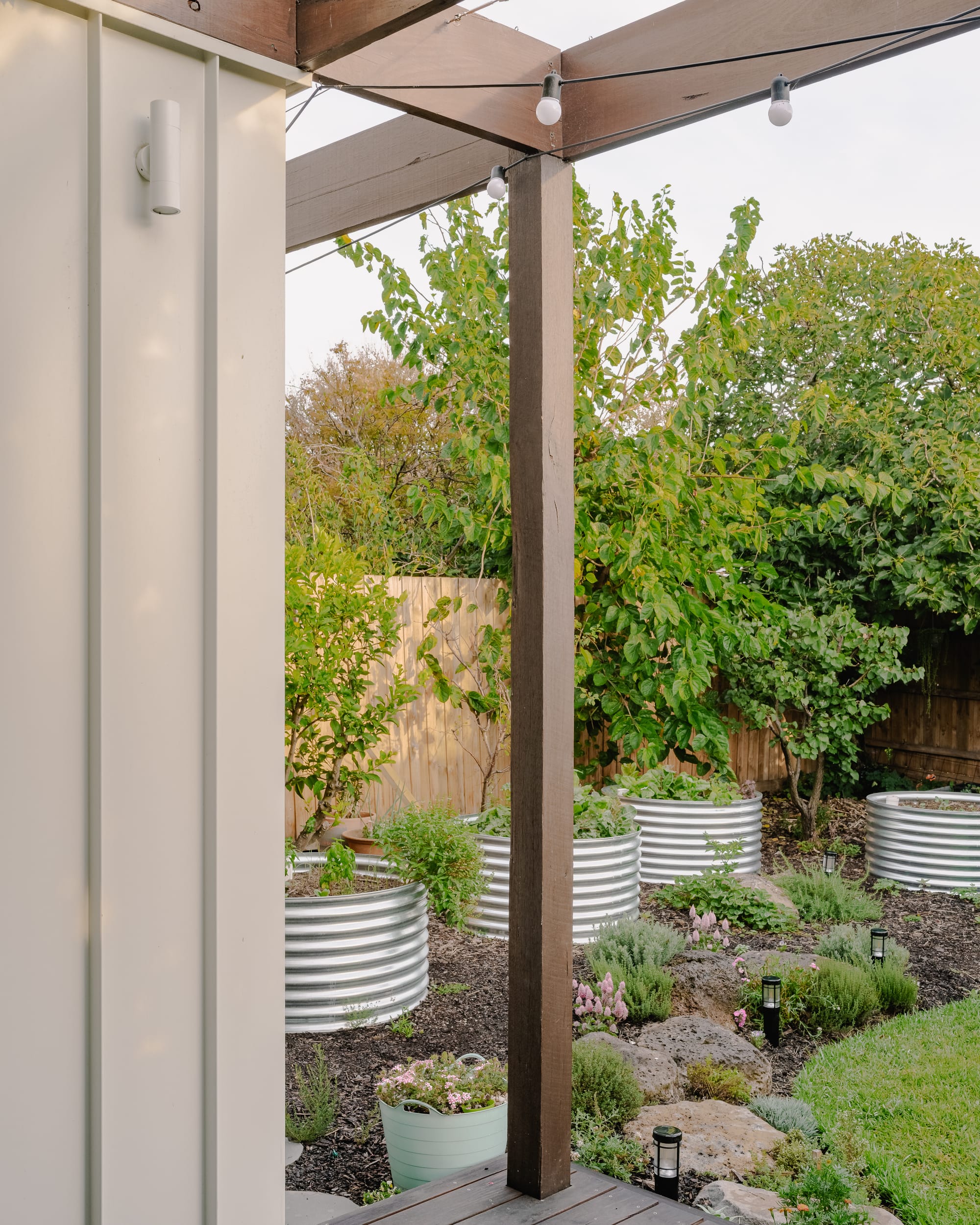 House in Preston by Ashley O'Neill Architects. Photography by Victor Vieaux. Timber pole in centre of image, attached to timber pergola from above and timber deck from below, in front of a residential garden with tin planters, rocks, mulch, grass and pink flowers. 