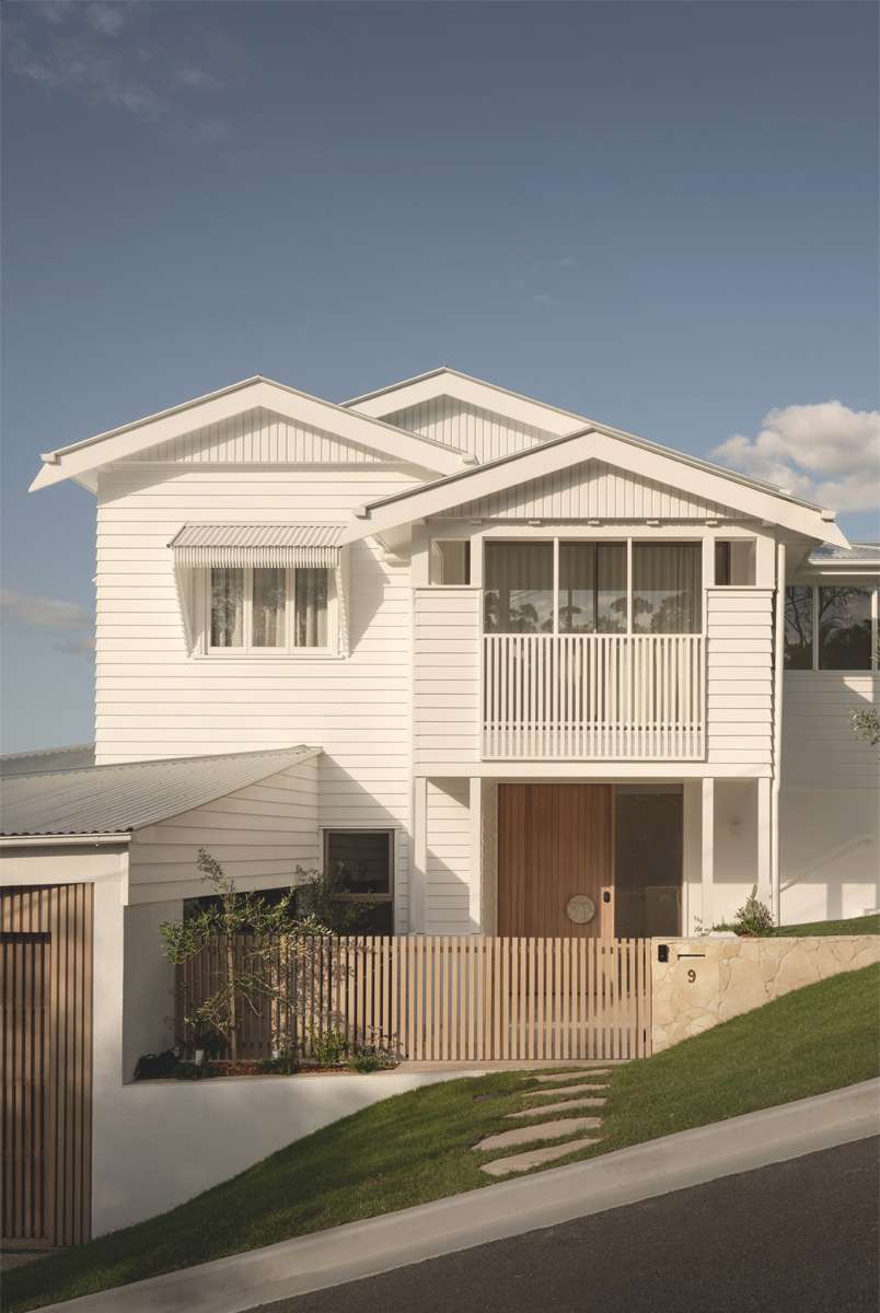 Paddington Queenslander by Kelder Architects. Photography by Kemp Photography. Street view of two storey white timber clad Queenslander on steep road, with green grass, stone pavers leading to a timber and stone fence and a large timber front door.