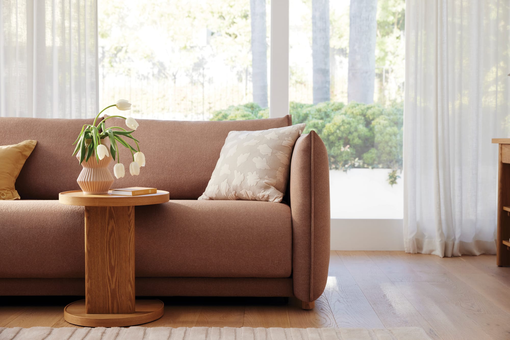 Byron Sofa Bed in Blush Sunset by Koala. Terracotta sofa with timber coffee table, white flowers in a vase and white sheer curtains in the background in front of window.