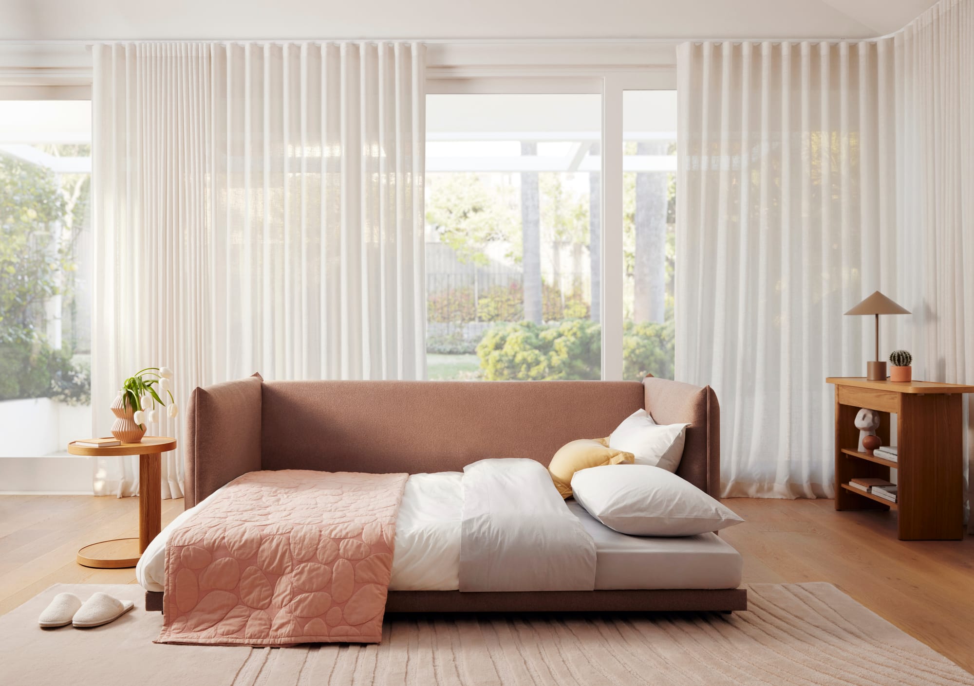 Byron Sofa Bed in Blush Sunset by Koala. Terracotta fold out sofa bed with coral throw rug in room with timber flooring, white sheer curtains and timber furnishings.