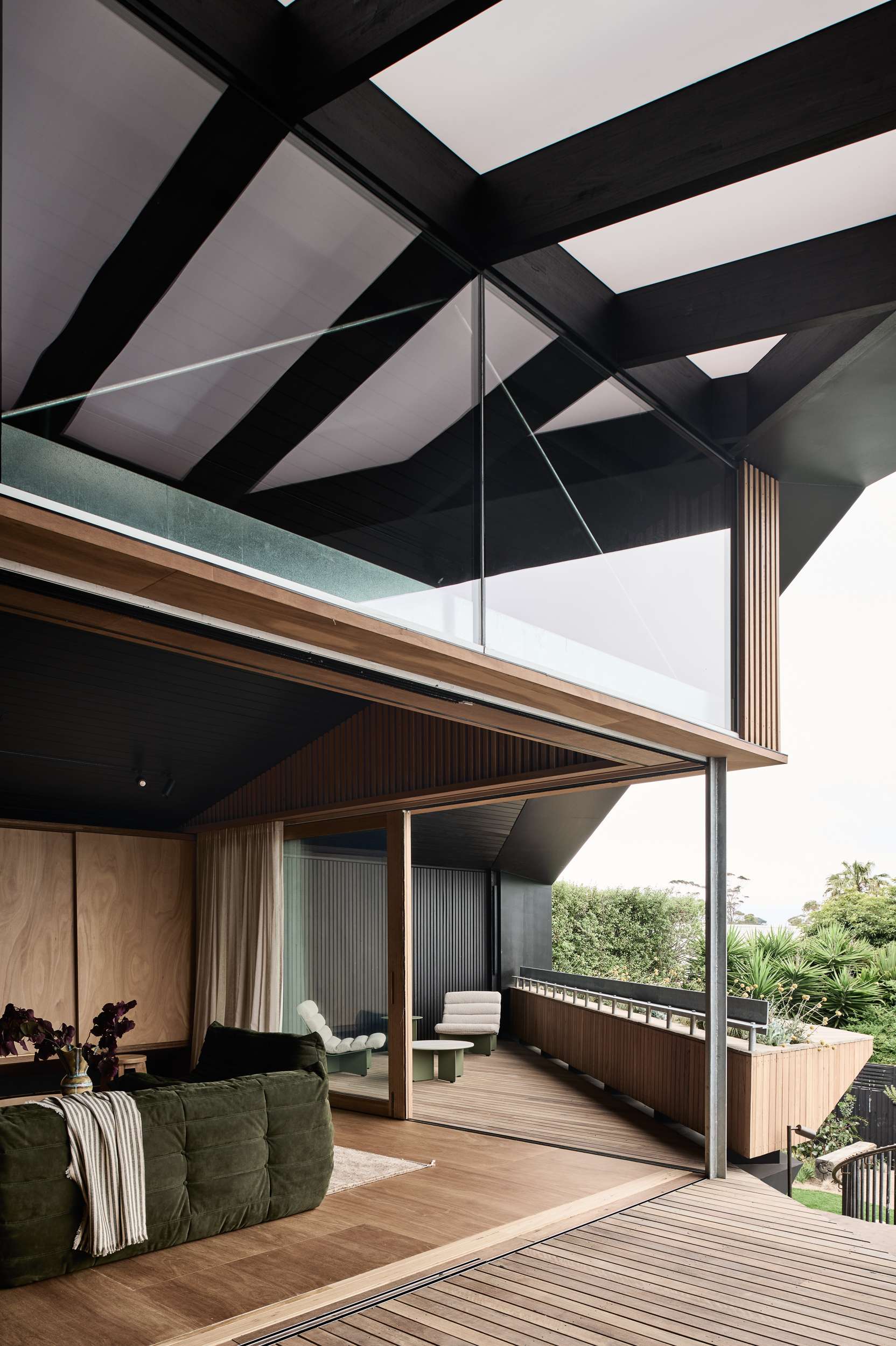 Kross House by FIGR Architecture. Photography by Tom Blachford. Open air interior and exterior timber clad entertaining areas, with a dark green velvet couch in the living area and green and white outdoor armchairs on the balcony.
