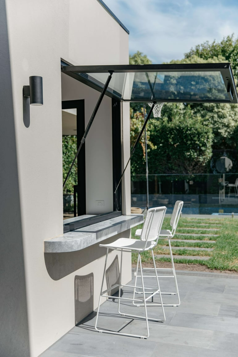 Little Moffat by Merrylees Architecture. Photography by Siy Studio. Servey window opening onto backyard with small concrete floating bench and white chairs on tiled patio with grass and pool in background. 