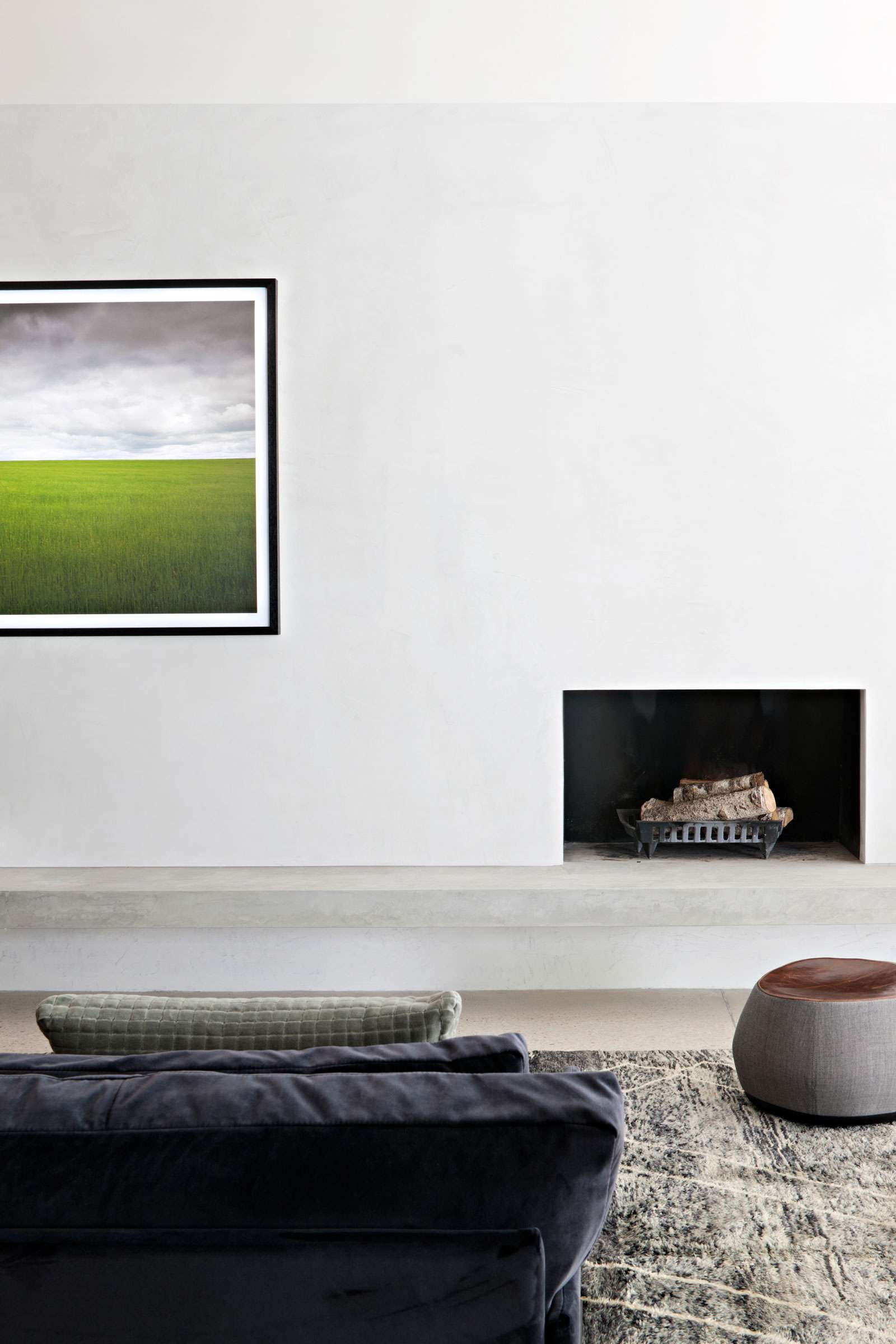 Victoria Gardens Home by Lucy Clemenger Architects. Photography by Shannon McGrath. Minimal living space with concrete and limewashed floating fireplace and wall, navy velvet couch in foreground and landscape imagery on wall.