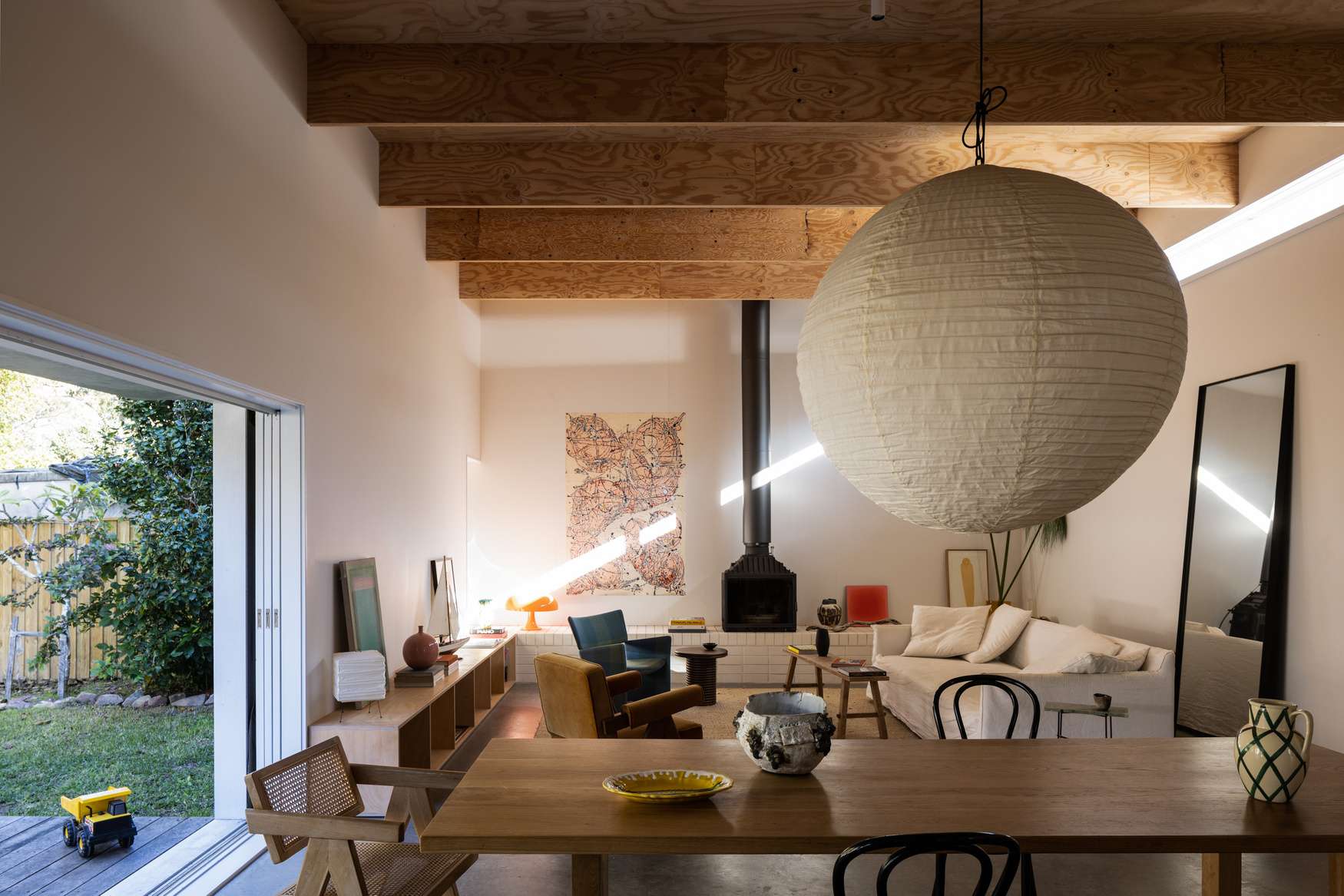 Drip Dry House by Marker Architecture. Photography by Simon Whitebread. Open plan living and dining space opening onto a timber deck and garden, with exposed timber beams, pendant lighting and a black fireplace inside.