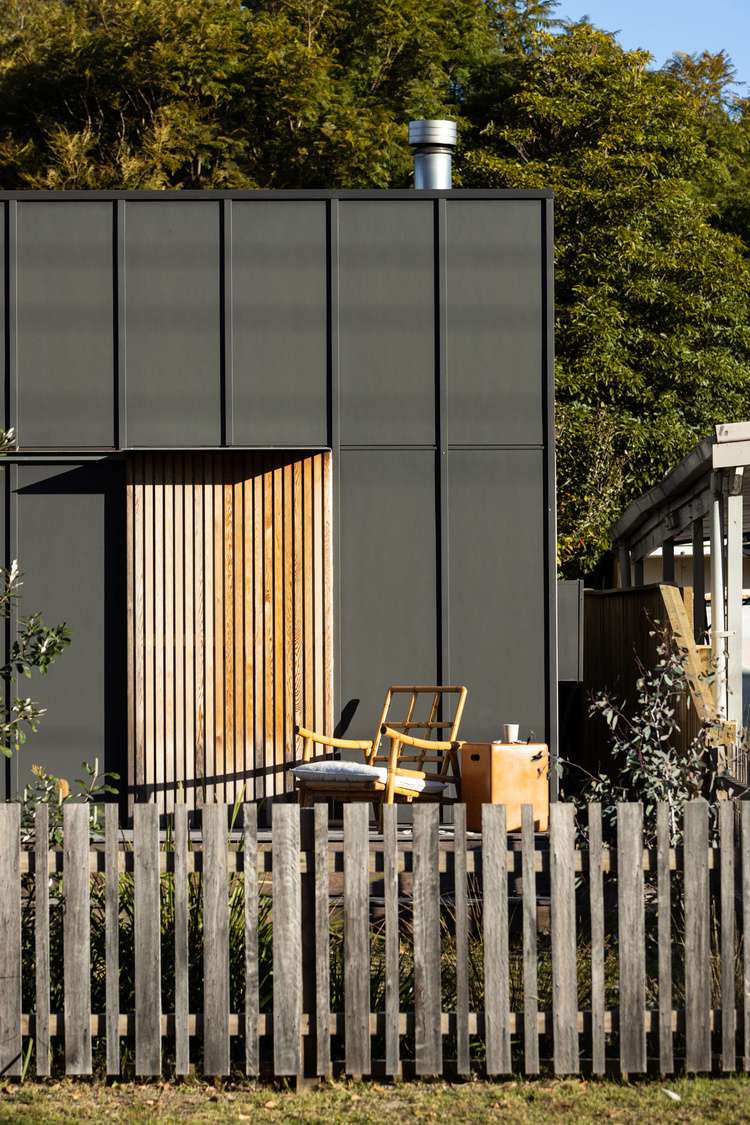 Drip Dry House by Marker Architecture. Photography by Simon Whitebread. Facade of black timber panelled exterior of home, with reclaimed timber clad door, worn wood picket fence and retro bamboo arm chair.