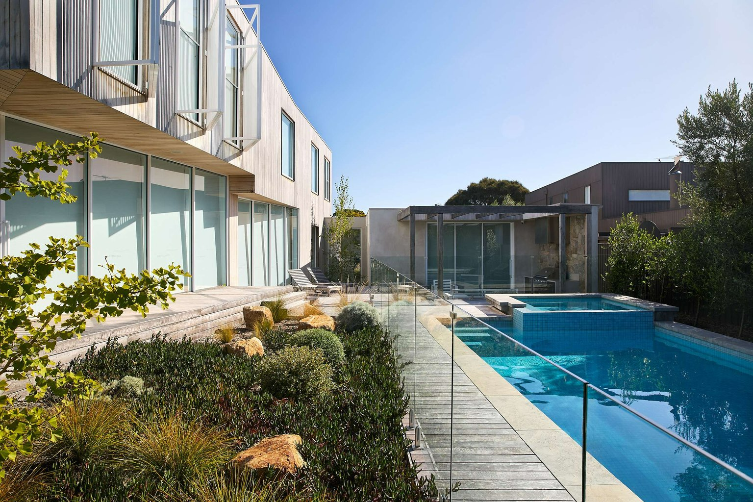 Peninsula House by Merrylees Architecture. Photography by Erik Holt. Rear of residential property ith glass fenced pool to the left, weathered timber deck wrapping around home, pool house in right hand background and double storey white home to the left. 