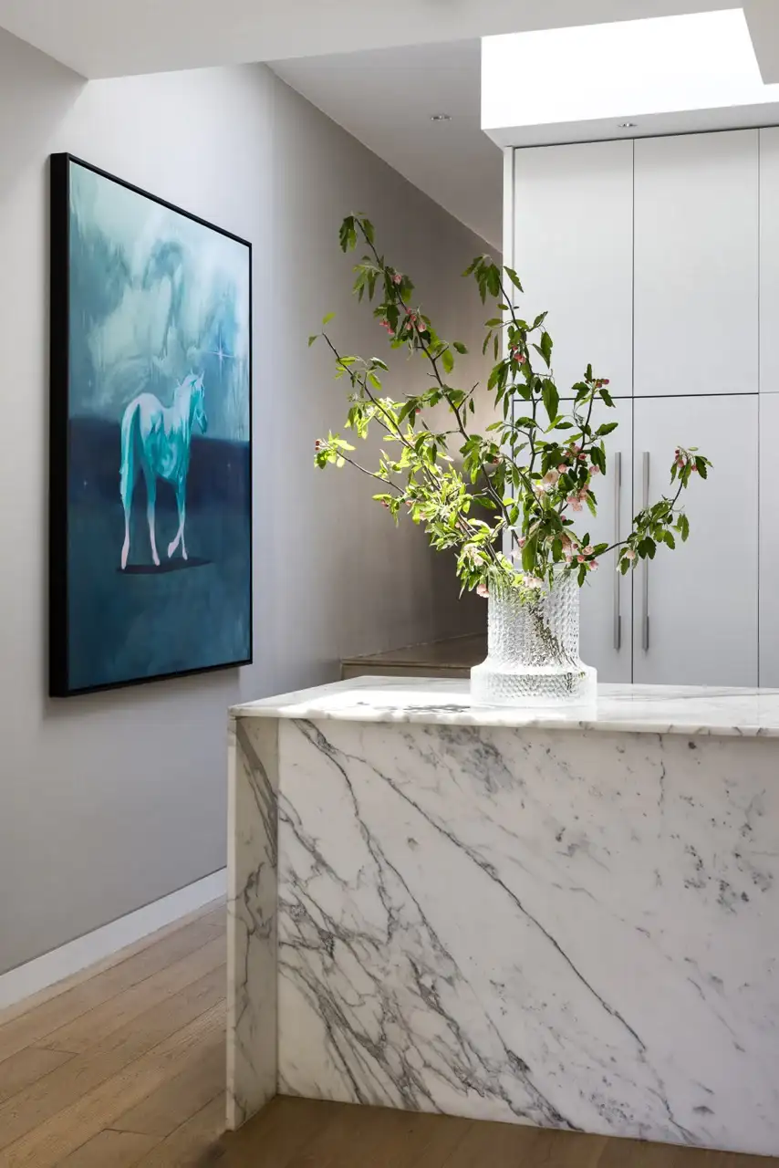 Paddington Terrace by FURNISHD. Photography by Dylan James. Kitchen with large marble island bench, crystal vase with white flowers, timber floors and blue wall art.