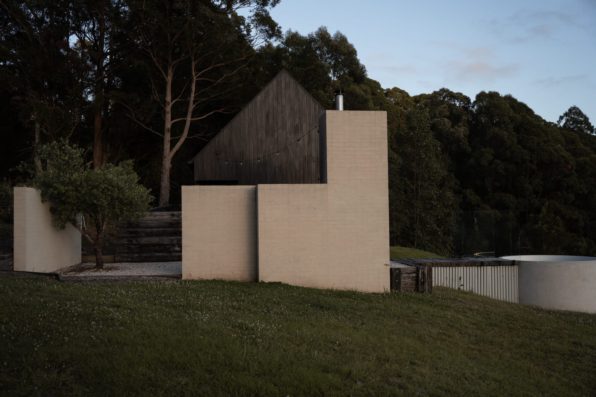 Round Hill Retreat by Round Hill Projects x SI.DE Architecture. Photography by In Sitū Studio. Cabon on green grassy hill with cream brick walls, black timber clad exterior with pitched roof and dense forest in background.