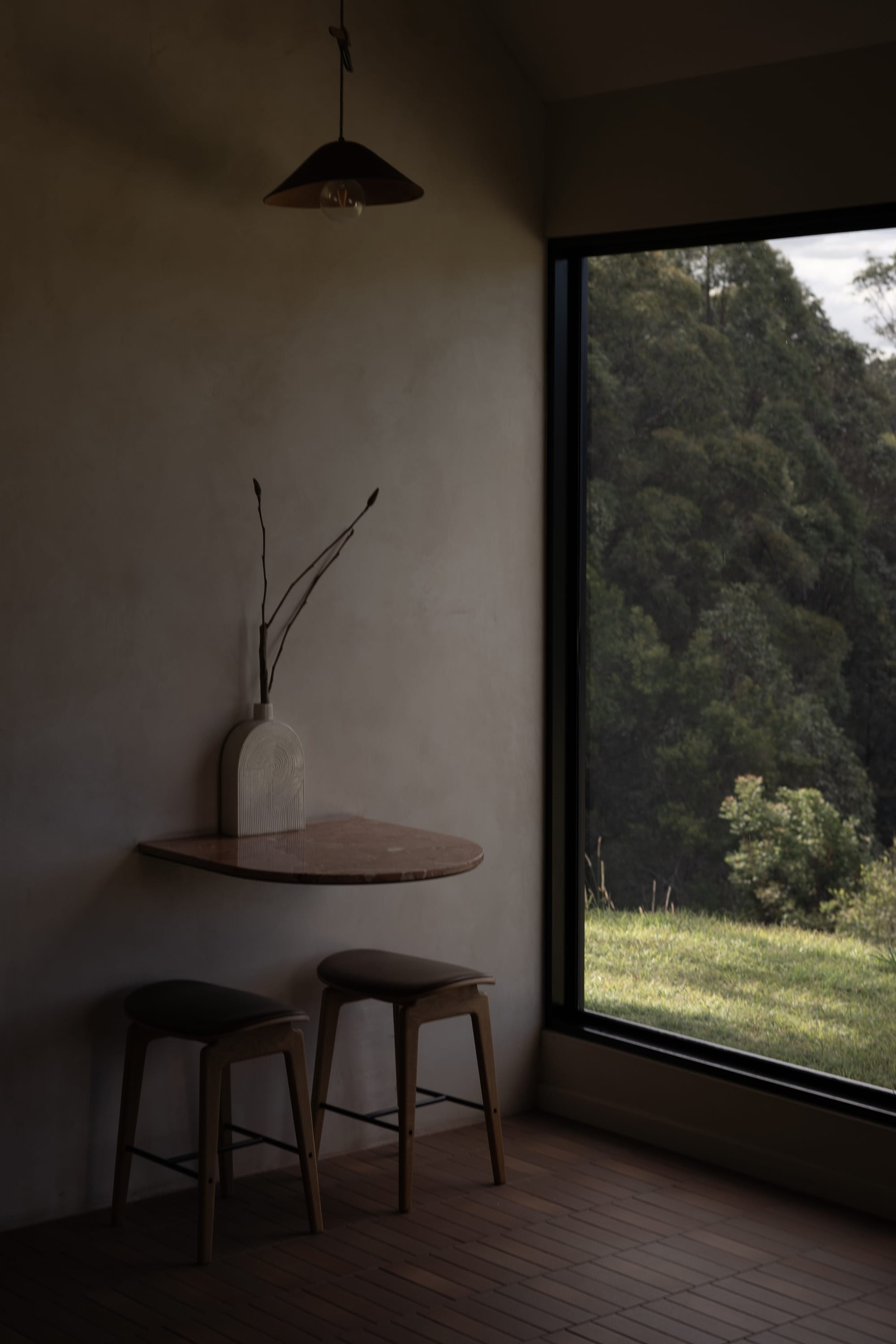 Round Hill Retreat by Round Hill Projects x SI.DE Architecture. Photography by In Sitū Studio. Floating stone tabletop with two timber stools in room with terracotta floortiles, lime washed walls and full height black framed windows overlooking green hillside and forest.