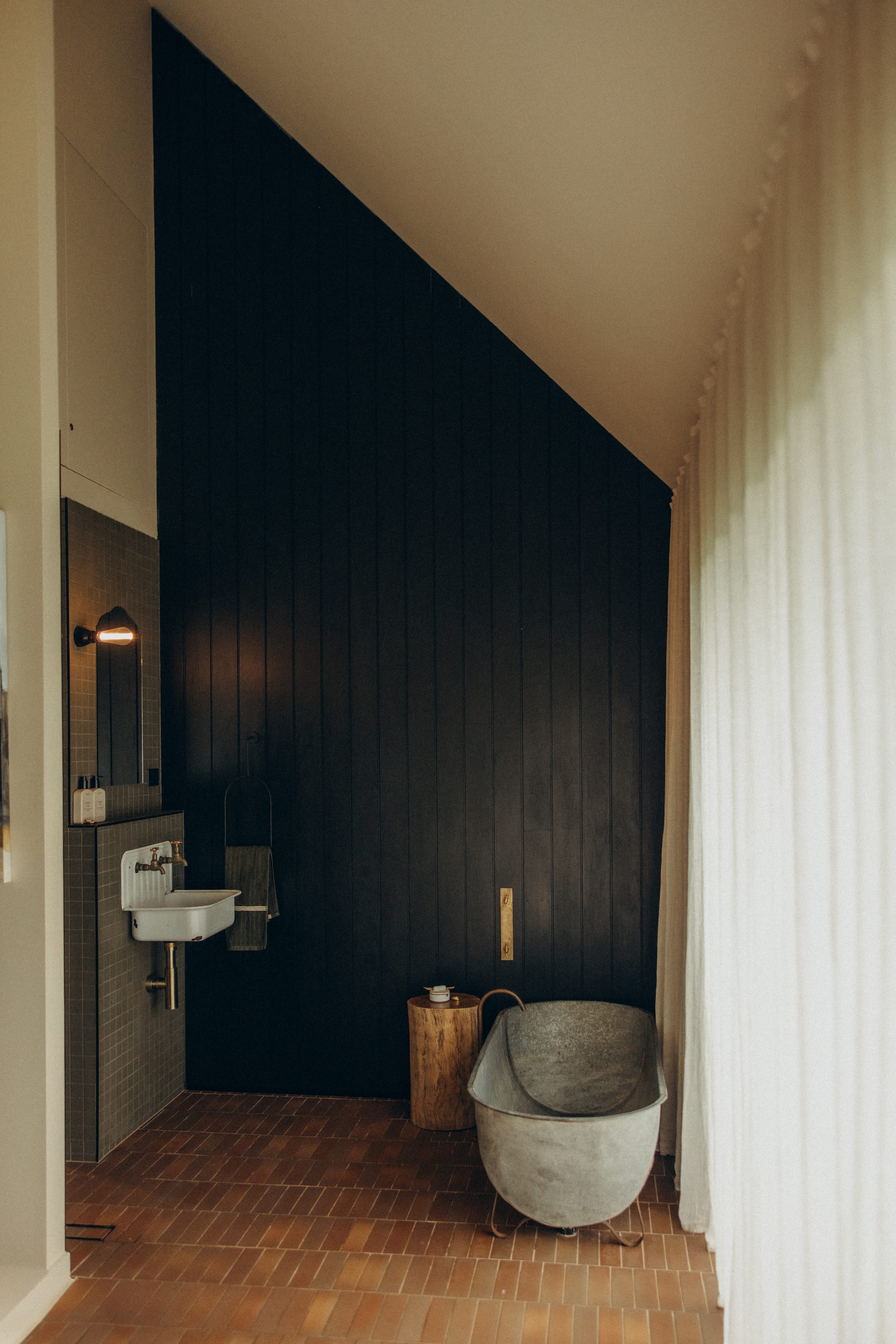 Round Hill Retreat by Round Hill Projects x SI.DE Architecture. Photography by Nicci K. Bathroom with pitched ceiling, black timber clad walls, terracotta floor tiles, white sheer curtains to the left and a tin bathtub with a timber side table.