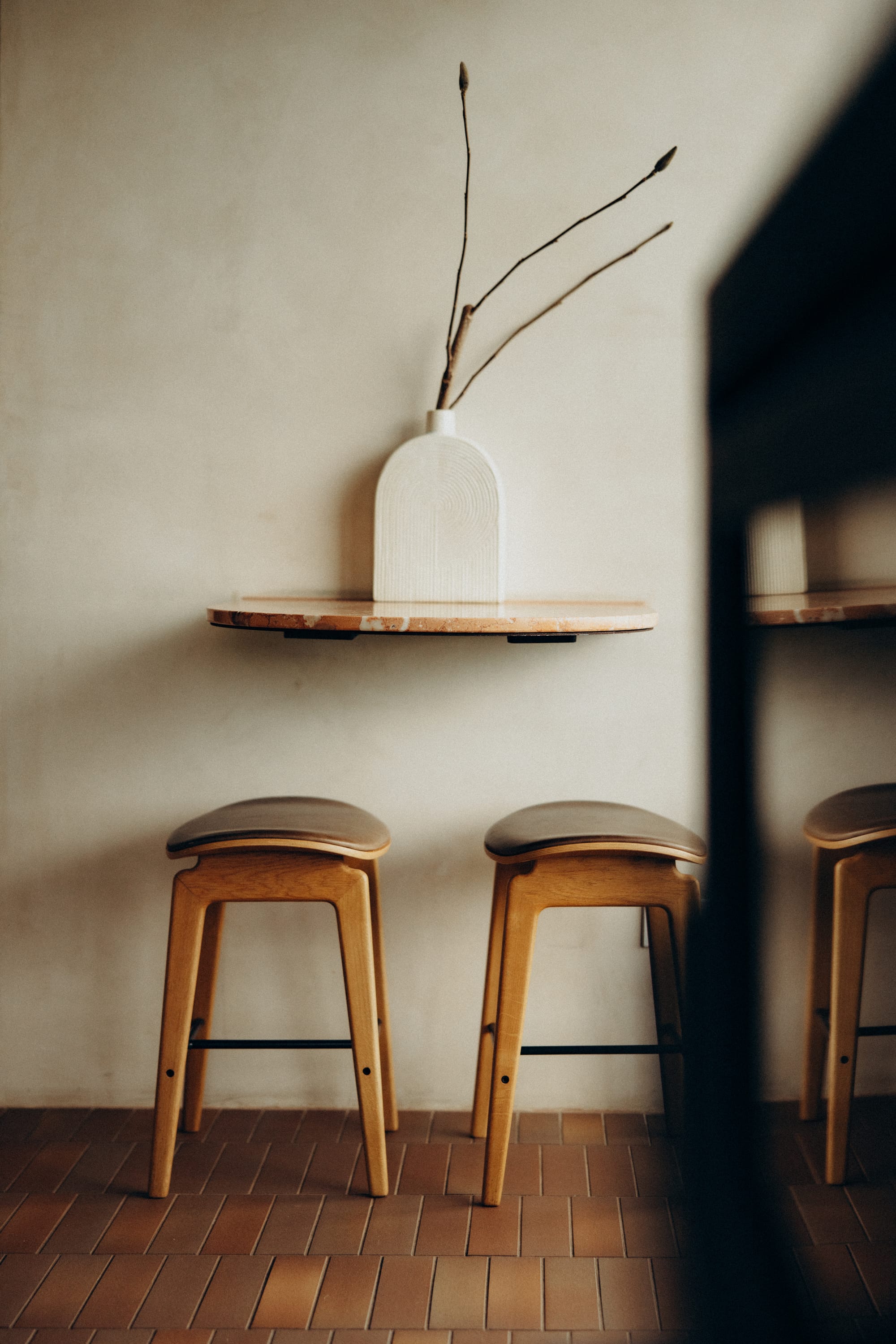 Round Hill Retreat by Round Hill Projects x SI.DE Architecture. Photography by Nicci K. Timber and cream leather stools in front of limewashed wall with floating half-circle shelf with large white vase, on terracotta floor tiles.