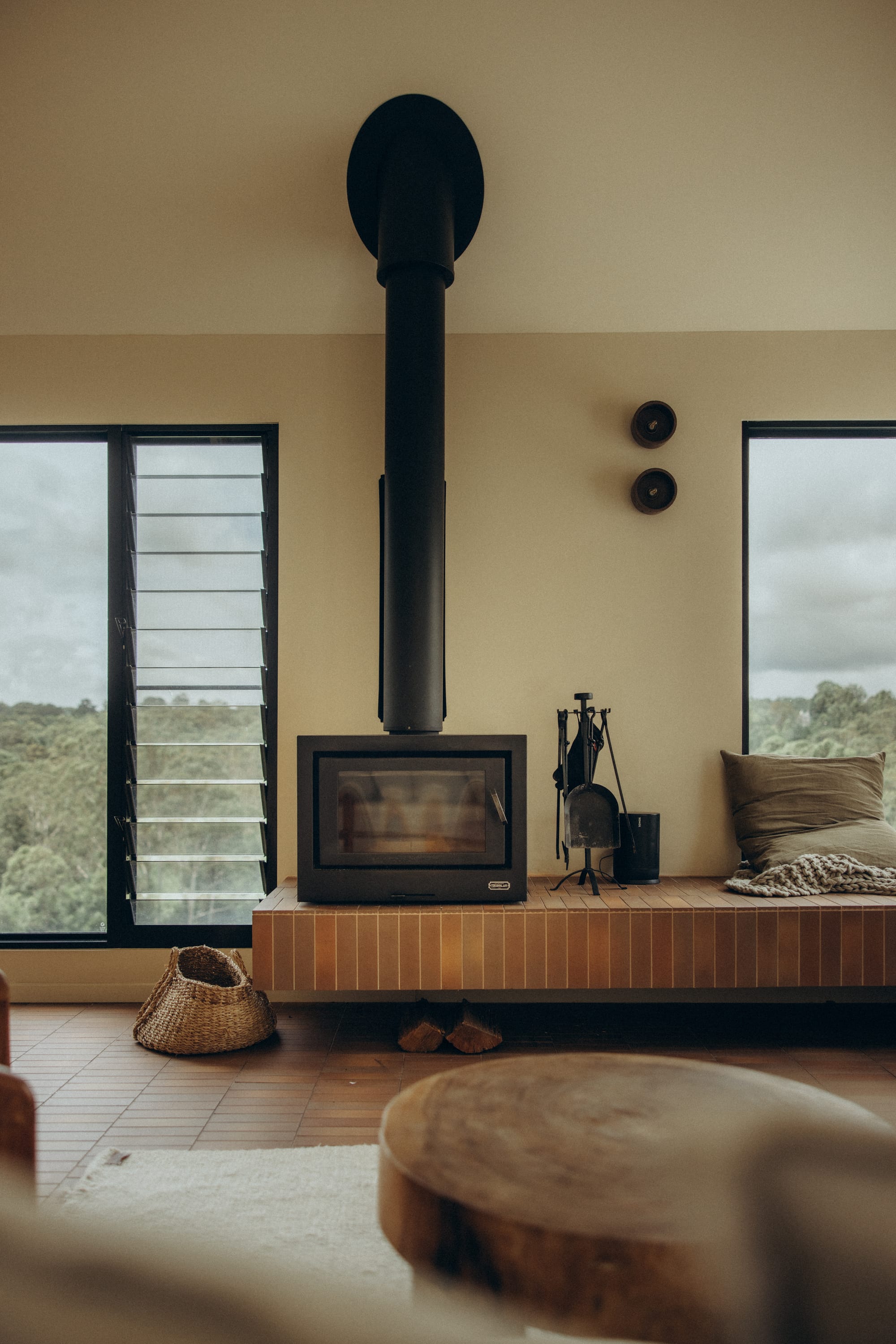 Round Hill Retreat by Round Hill Projects x SI.DE Architecture. Photography by Nicci K. Living room with black fireplace on floating tiled bench, with limewashed walls, terracotta floor tiles, louvred windows and a round timber coffee table.