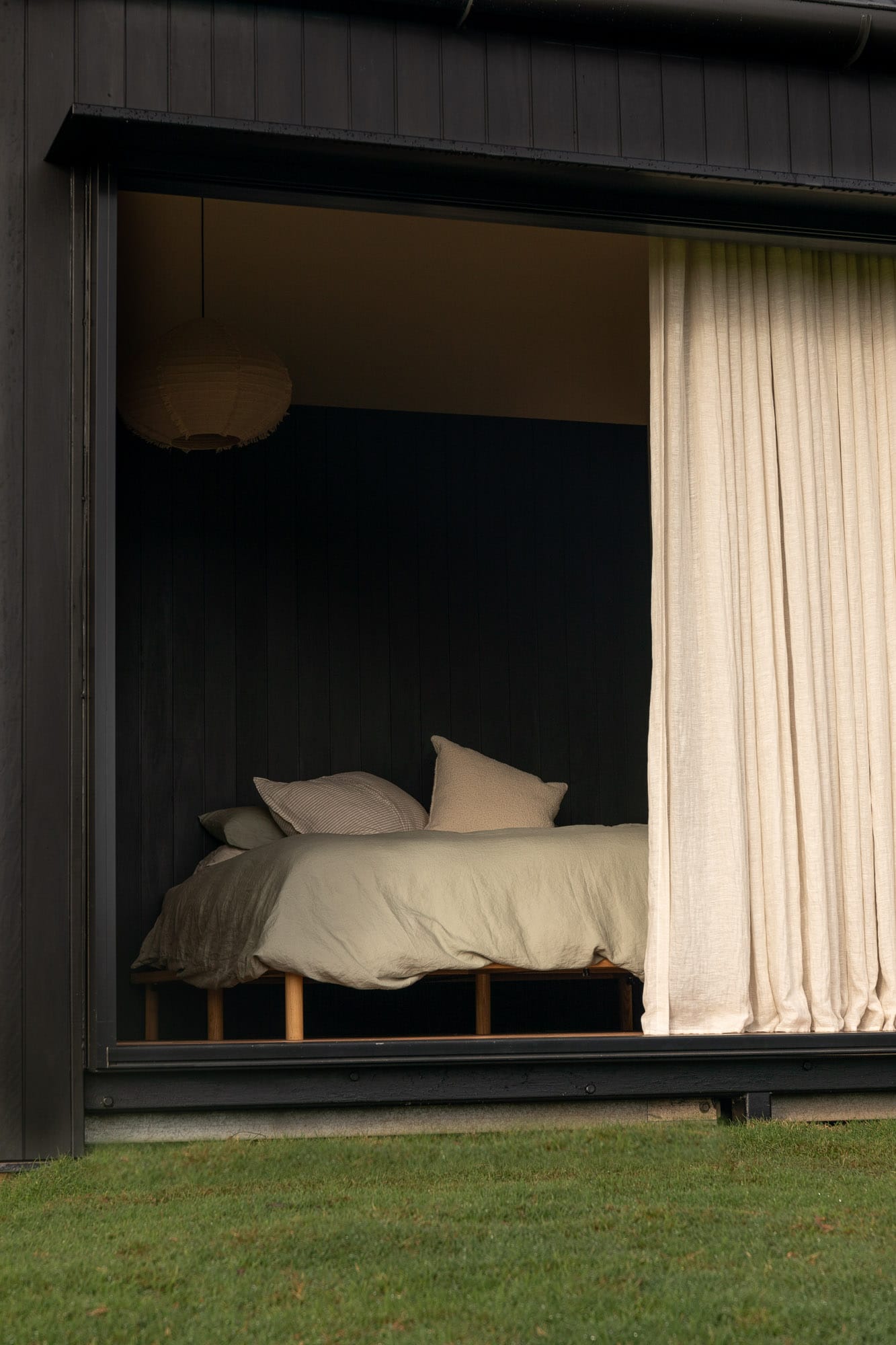 Round Hill Retreat by Round Hill Projects x SI.DE Architecture. Photography by Tim Birch. Bedroom shot through full height window opening on timber clad exterior, with linen curtains and light green bed linen.