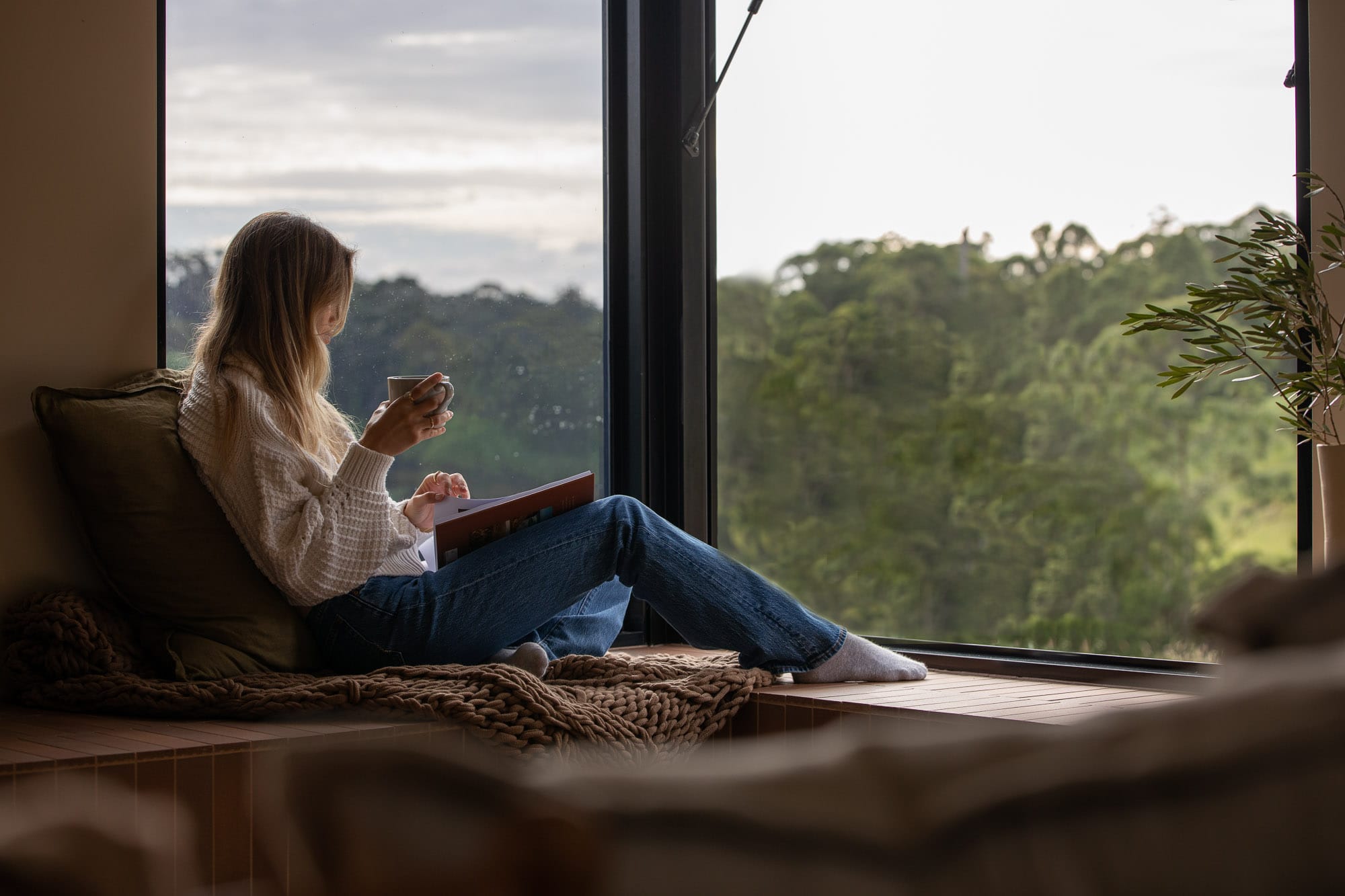 Round Hill Retreat by Round Hill Projects x SI.DE Architecture. Photography by Tim Birch. Woman sitting with a cup of tea and a book on a tiled bench seat in front of full height windows overlooking dense treeline.