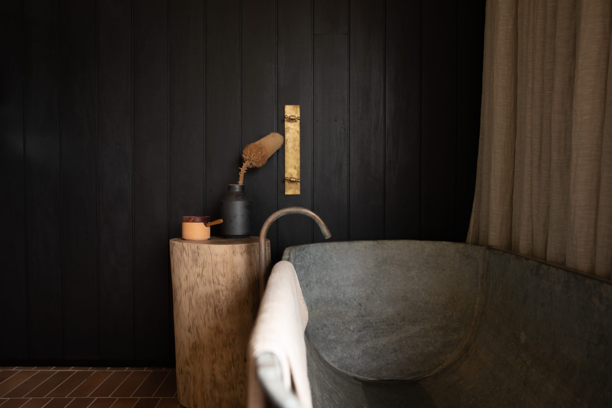 Round Hill Retreat by Round Hill Projects x SI.DE Architecture. Photography by Tim Birch. Vintage tin bathtub in bathroom with terracotta floor tiles, black timber clad walls, beige linen sheers and a rustic timber side table.