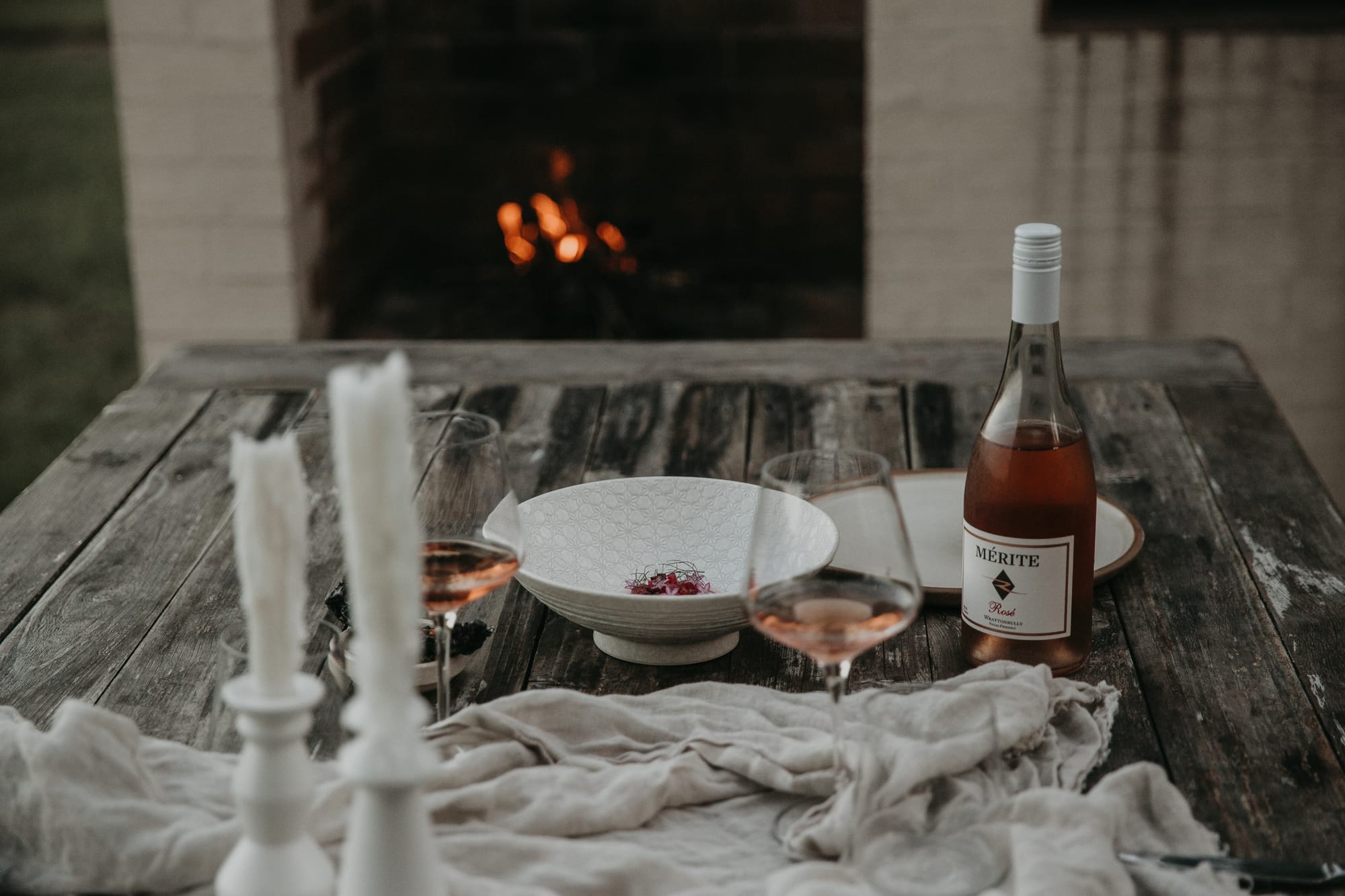 Round Hill Retreat by Round Hill Projects x SI.DE Architecture. Photography by Yani Rae. Weathered outdoor timber table, set with grey linen table covering, white plate, white candlesticks and pink wine with fireplace in the background