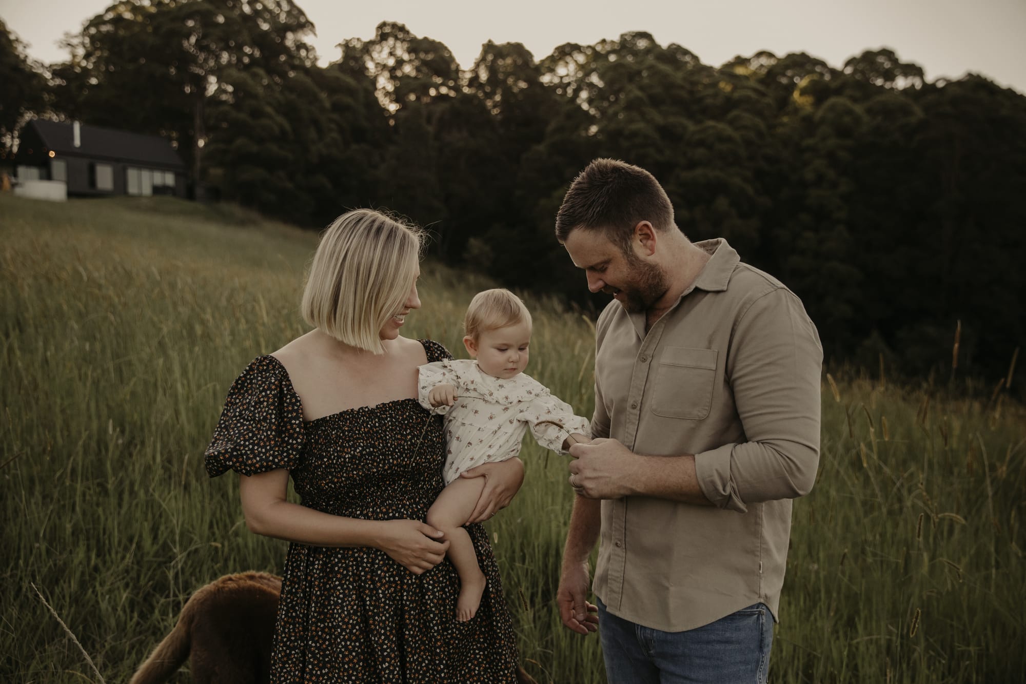 Alex and Chloe of Round Hill Projects. Photography by Yani Rae. A woman with blond hair holding a baby, standing next to a man in a beige shirt in a field, with a black cabin visible in the background.