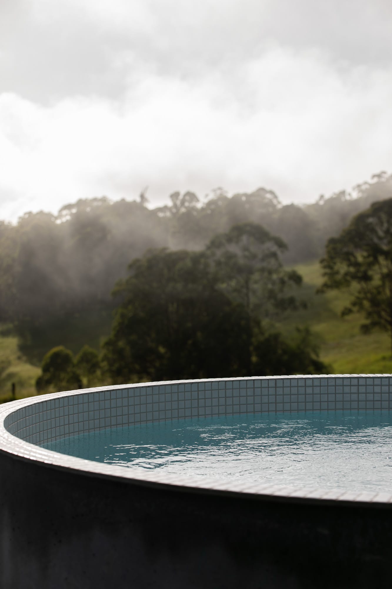 Round Hill Retreat by Round Hill Projects x SI.DE Architecture. Photography by Tim Birch. Round above ground tiled plunge pool with black exterior and white square tiles inside, overlooking rolling green hills, paddocks and forests.