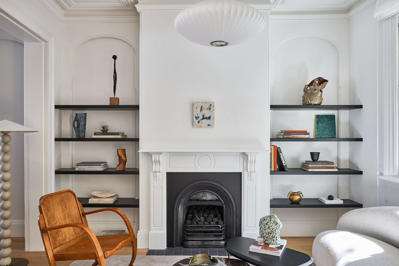 Crown St Terrace by Studio Arkive. Living room with white walls, original heritage fireplace and mantle, floating black shelving on either side, a timber rocking chair, white curved couch and a black coffee table. 
