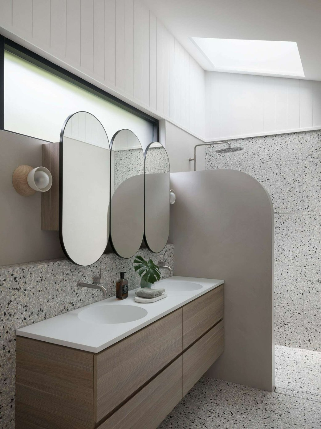 Palm 01 by Watershed Architects. Photography by Tom Ferguson. Bathroom with terrazzo floor and wall tiles, curved beige shower screen, three curved wall mirrors and timber cabinetry.