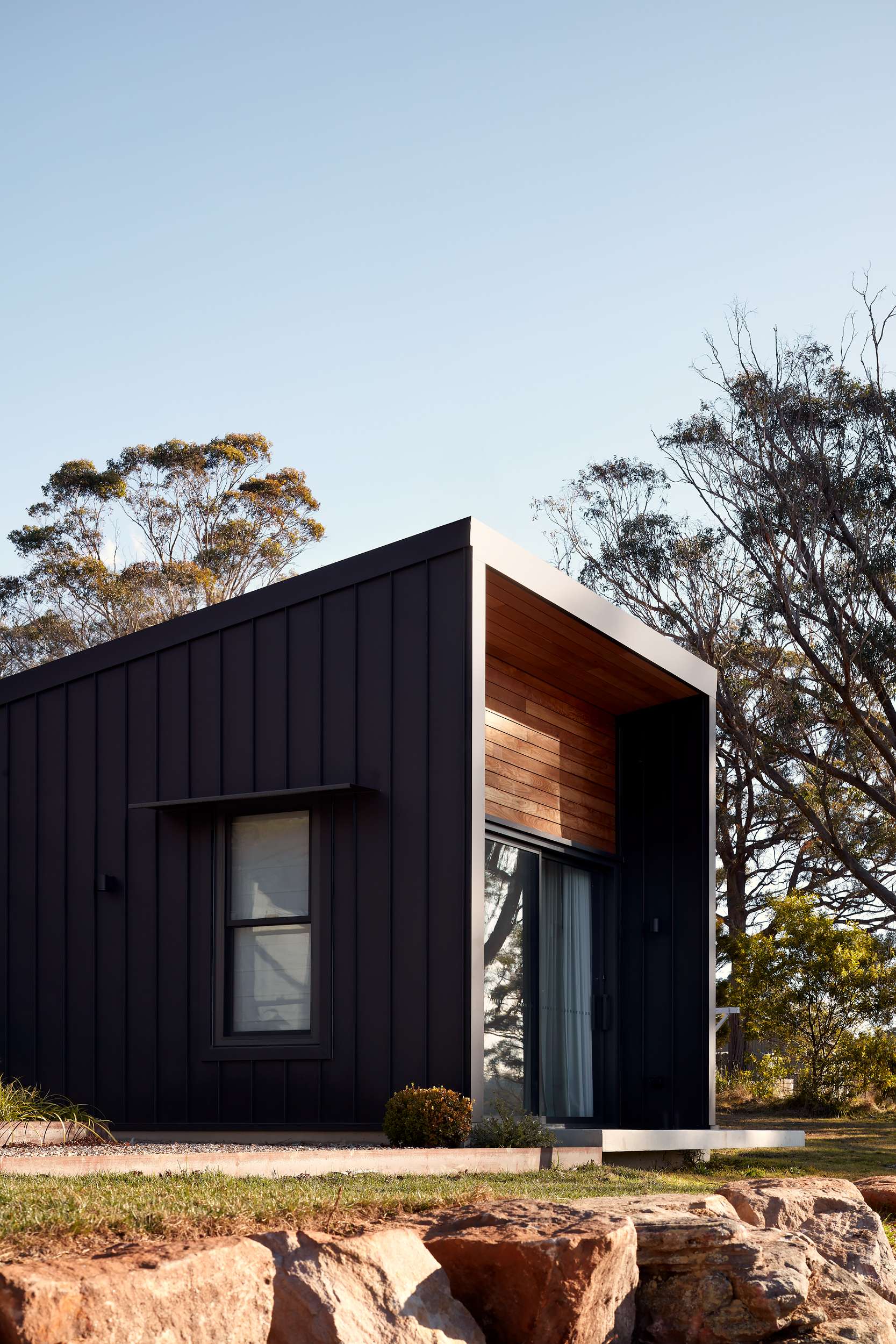 House Woodlands by AO Design Studio. Photography by Luc Remond. Rear facade of black and timber modern residential structure, surrounded by native bushland and rocks. 