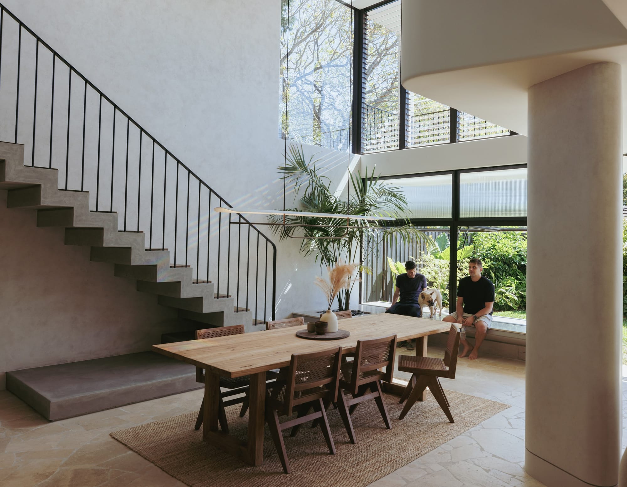 Little Birch by buck&simple. Photography by Tim Pascoe. Dining space with large timber table, concrete staircase with black railing and window seating in front of double storey black framed windows overlooking garden.