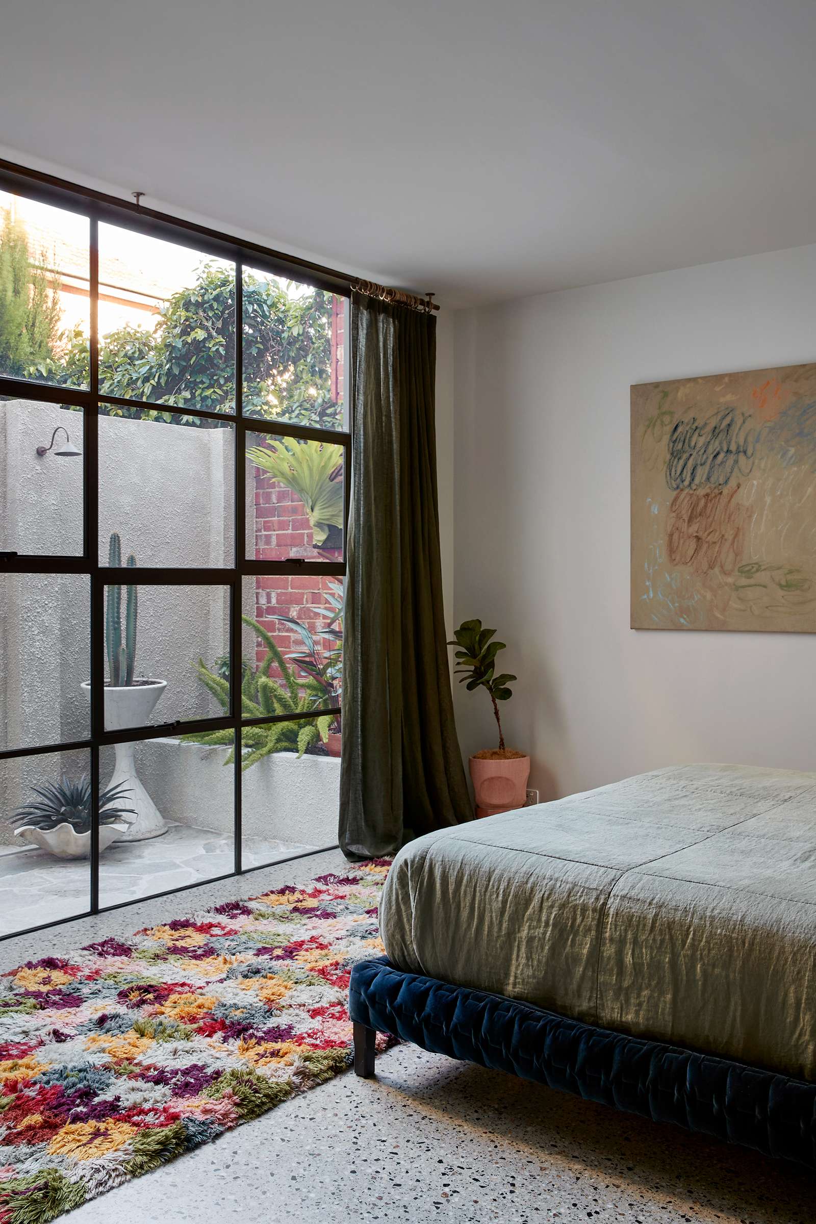 Fabrica by mcmahon and nerlich. Photography by Shannon McGrath. Bedroom with multicoloured tufted rug in front of black framed grid windows looking into paved courtyard with outdoor shower.