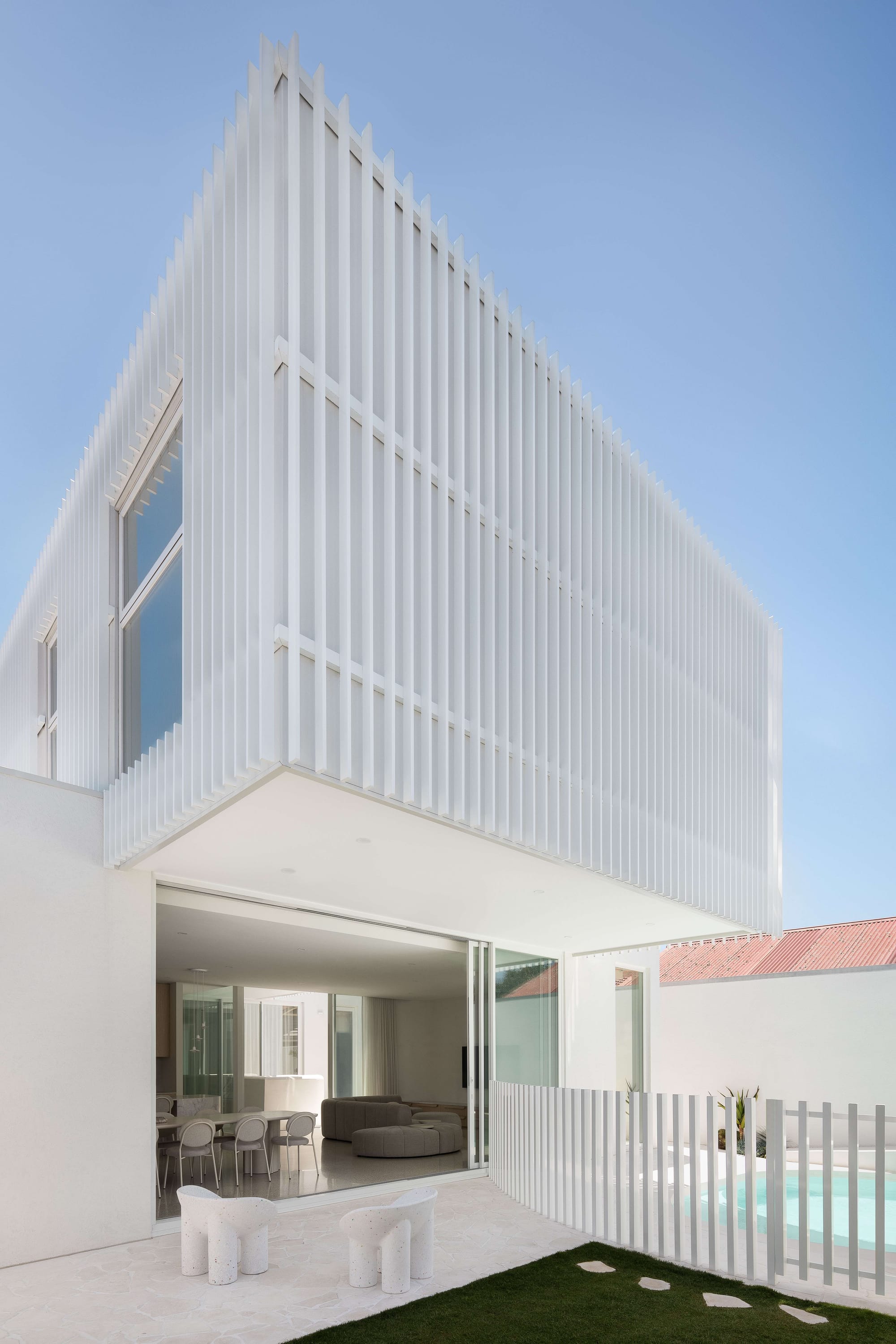 Alba House by Taouk Architects. Photography by Emily Bartlett. Rear facade of double storey home with pool, paved patio and overhanging second floor, with fluted white paneled exterior. 