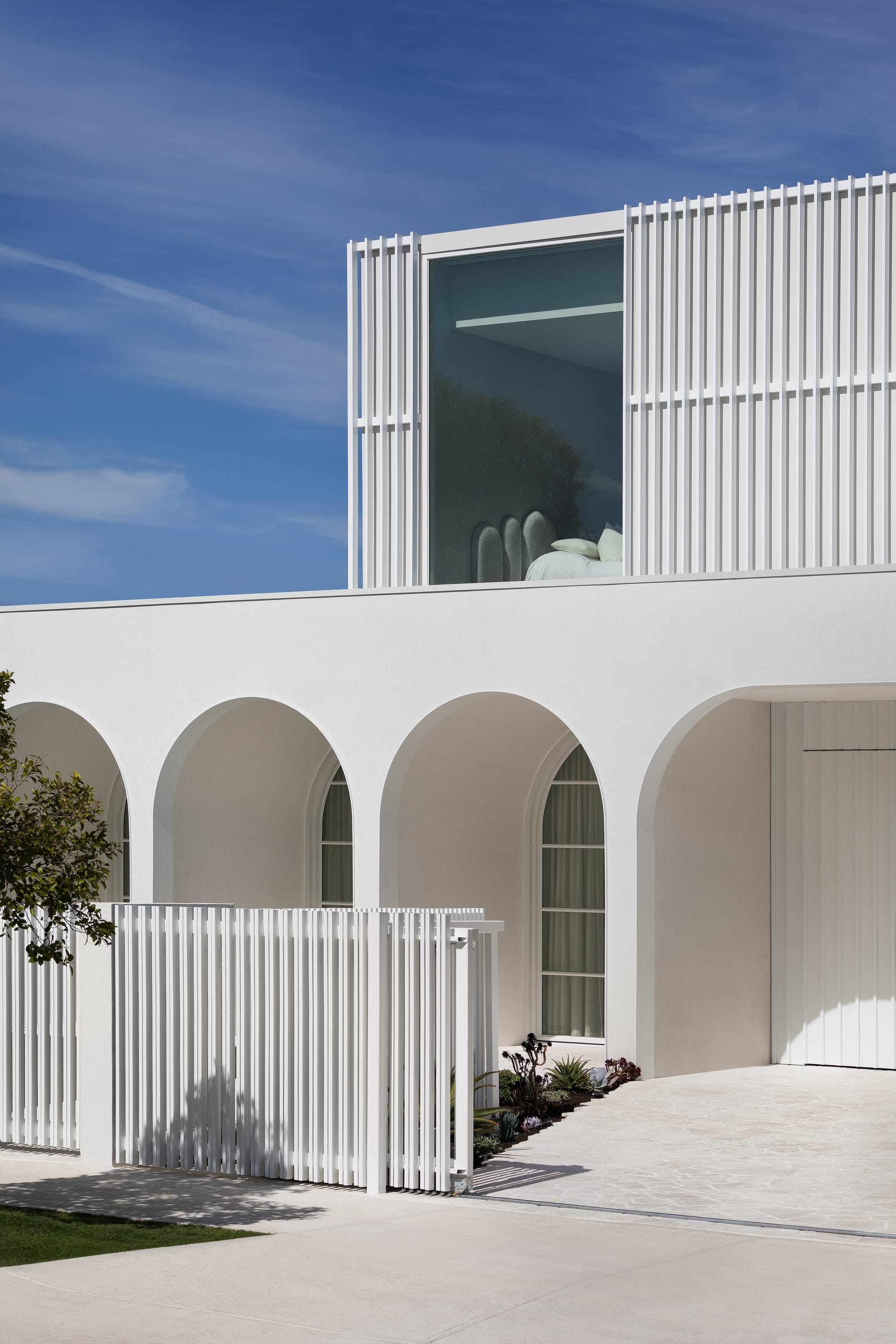 Alba House by Taouk Architects. Photography by Emily Bartlett. Rear facade of double storey home with pool, paved patio and overhanging second floor, with fluted white paneled exterior. 