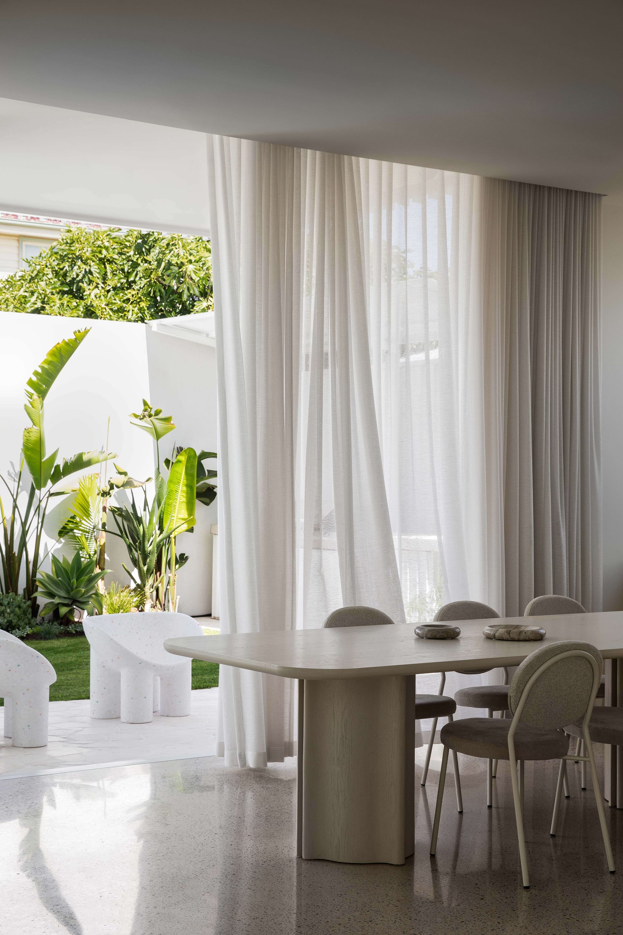 Alba House by Taouk Architects. Photography by Emily Bartlett. Dining space with large timber table, polished concrete floors, full hieght sliding glass doors with white sheer curtains opening onto backyard with white stone chairs and grass. 