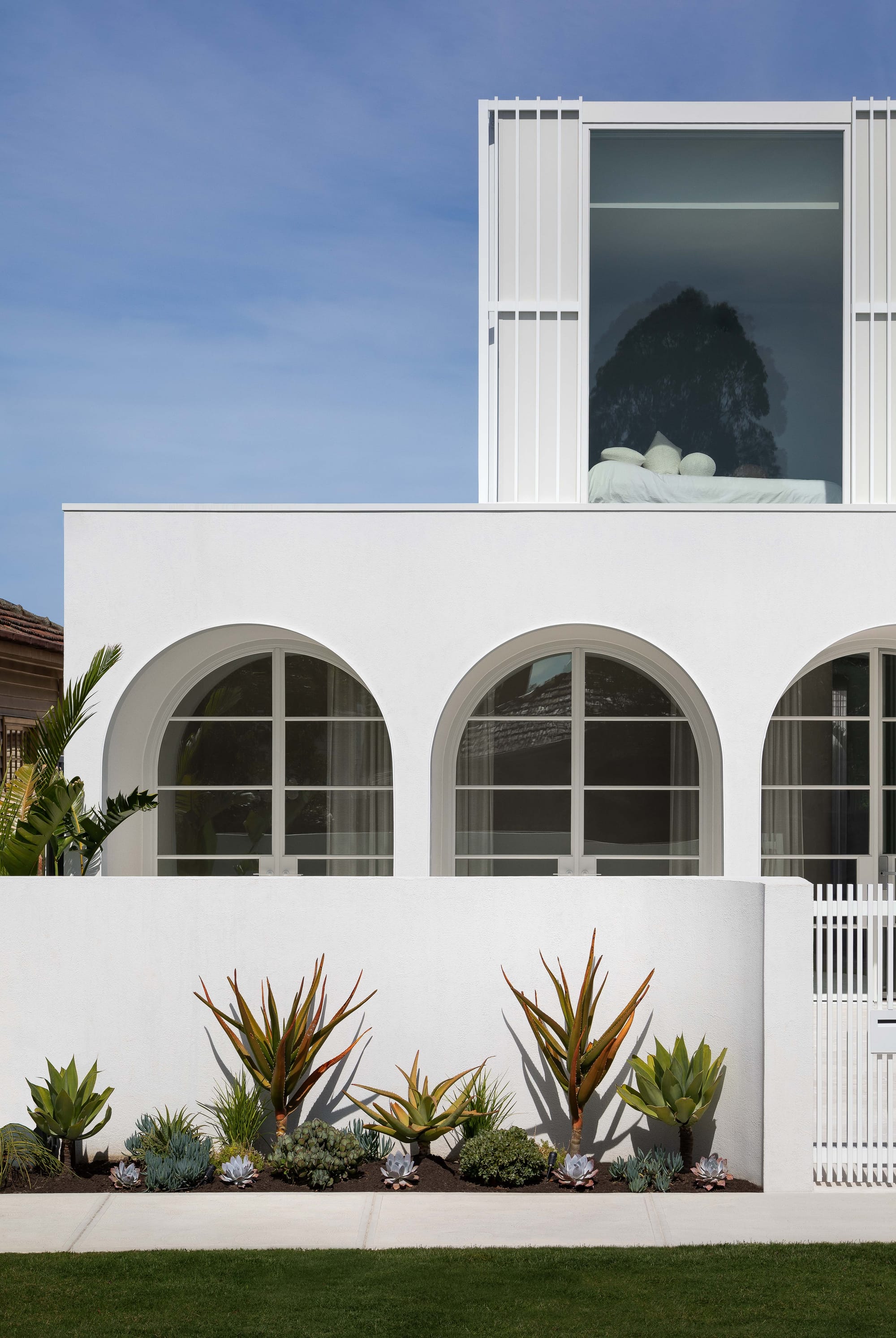 Alba House by Taouk Architects. Photography by Emily Bartlett. Facade of white double storey home in daylight with white rendered exterior, curved windows, and cactus garden alongside footpath. 