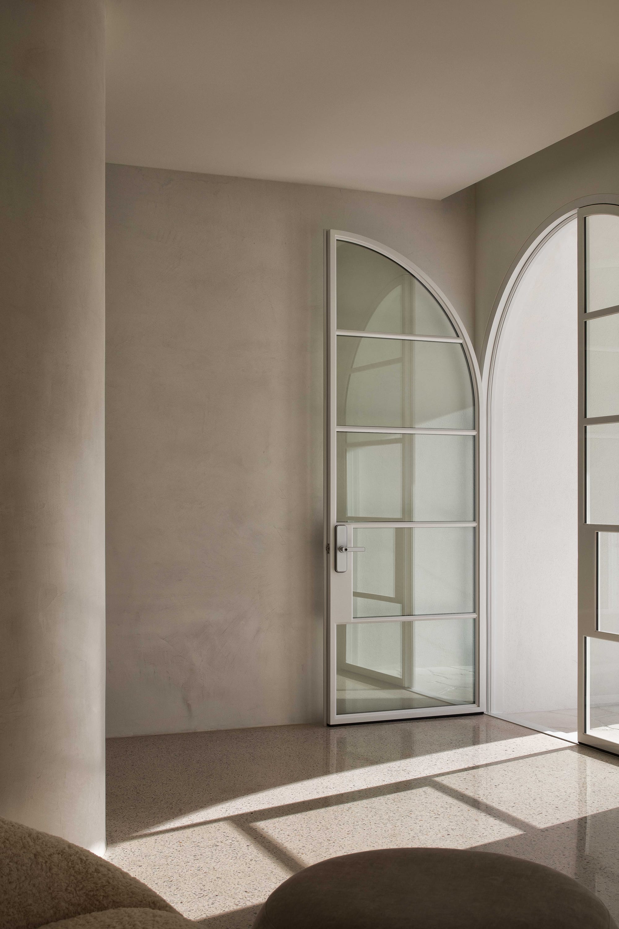 Alba House by Taouk Architects. Photography by Emily Bartlett. Space with limewashed walls, round pillars, polished concrete flooring and arched glass door letting light through space.