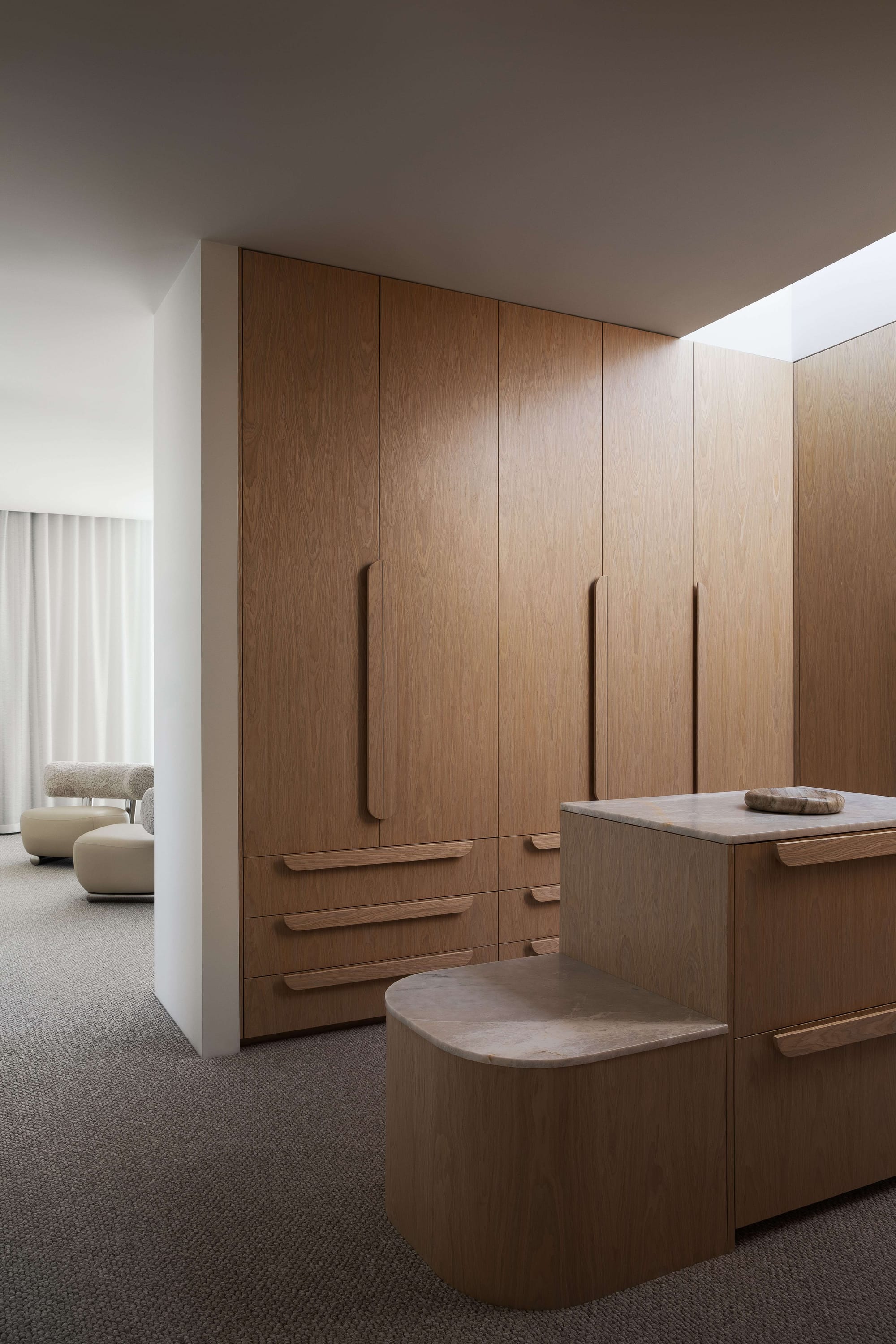Alba House by Taouk Architects. Photography by Emily Bartlett. Walk in wardrobe with grey carpeting, timber cabinetry with integrated timber handles and timber island with tiered stone benchtops.