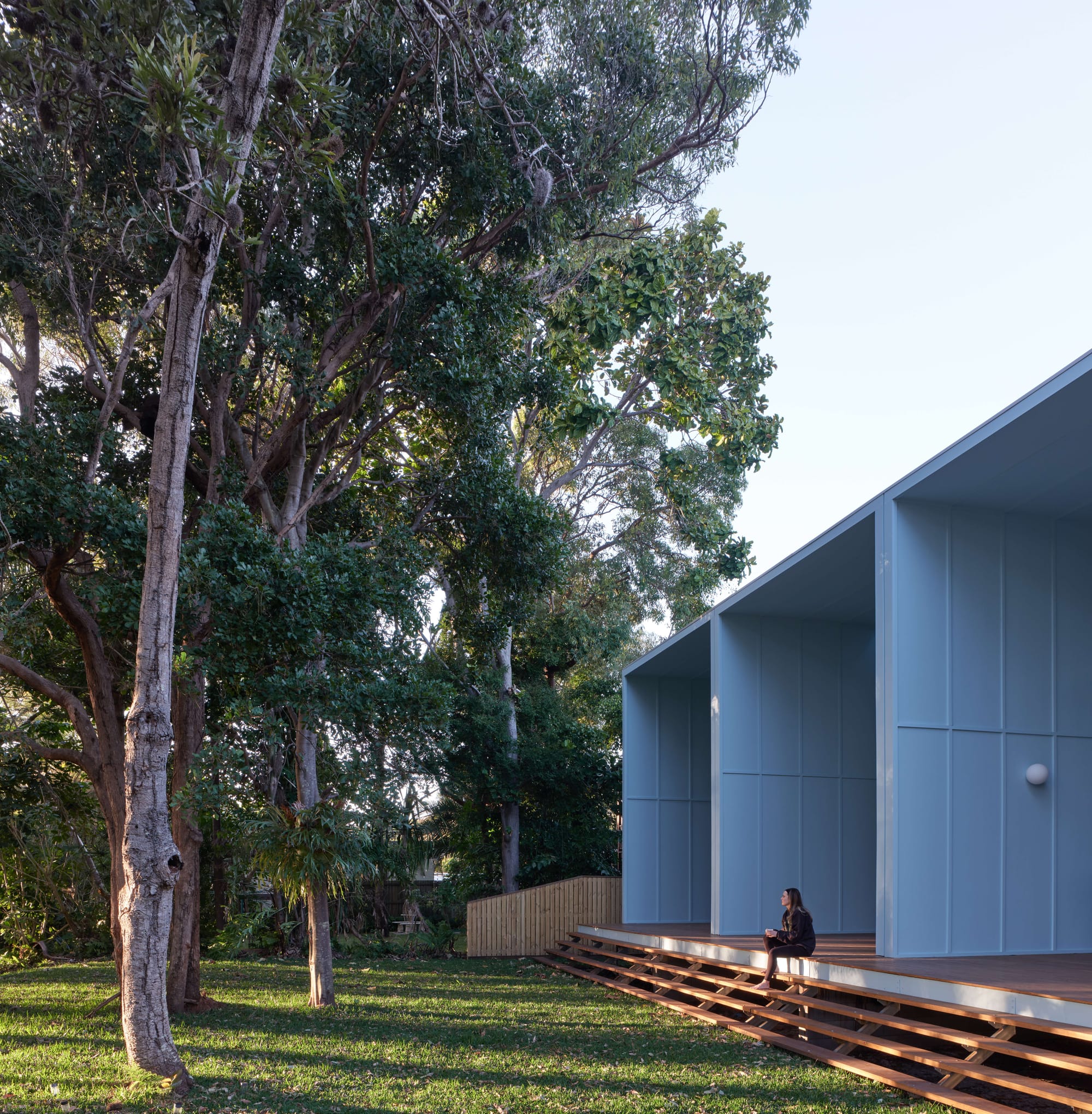 Blok Peregian by Blok Three Sisters. Photography by Christopher Frederick Jones. Rear facade of home with blue metal clad walls and a full-length timber deck with stairs leading to grassed backyard with native trees.
